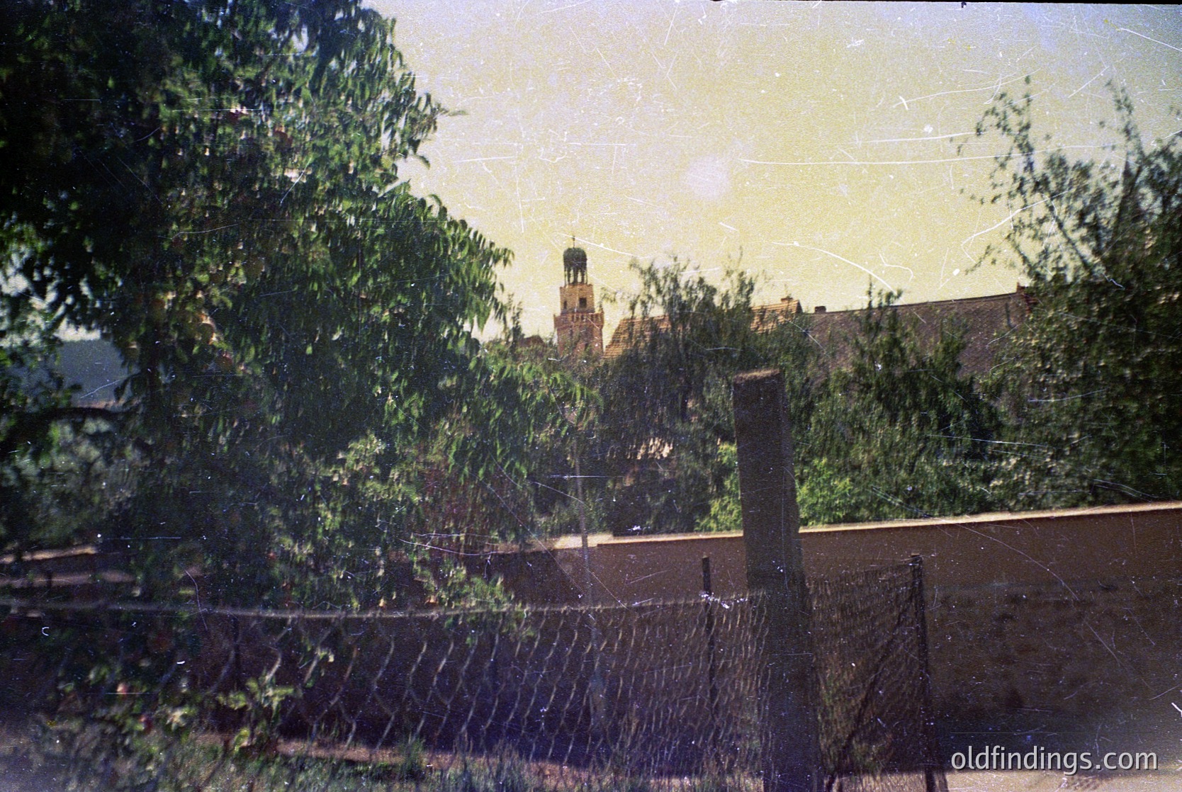 Vintage sepia-toned photo showing a distant church tower framed by foliage and a chain-link fence. Likely Eastern European urban setting, 1960s–1980s. Soft focus and slight film grain enhance nostalgic atmosphere.