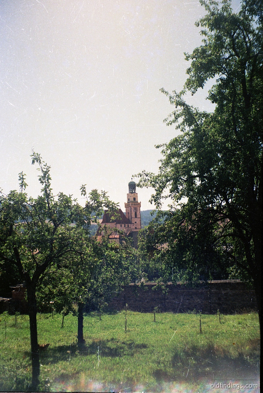 Historic brick church with tall bell tower framed by lush greenery and mature trees. Likely European countryside setting, possibly architecture. Stone wall and open field in foreground.