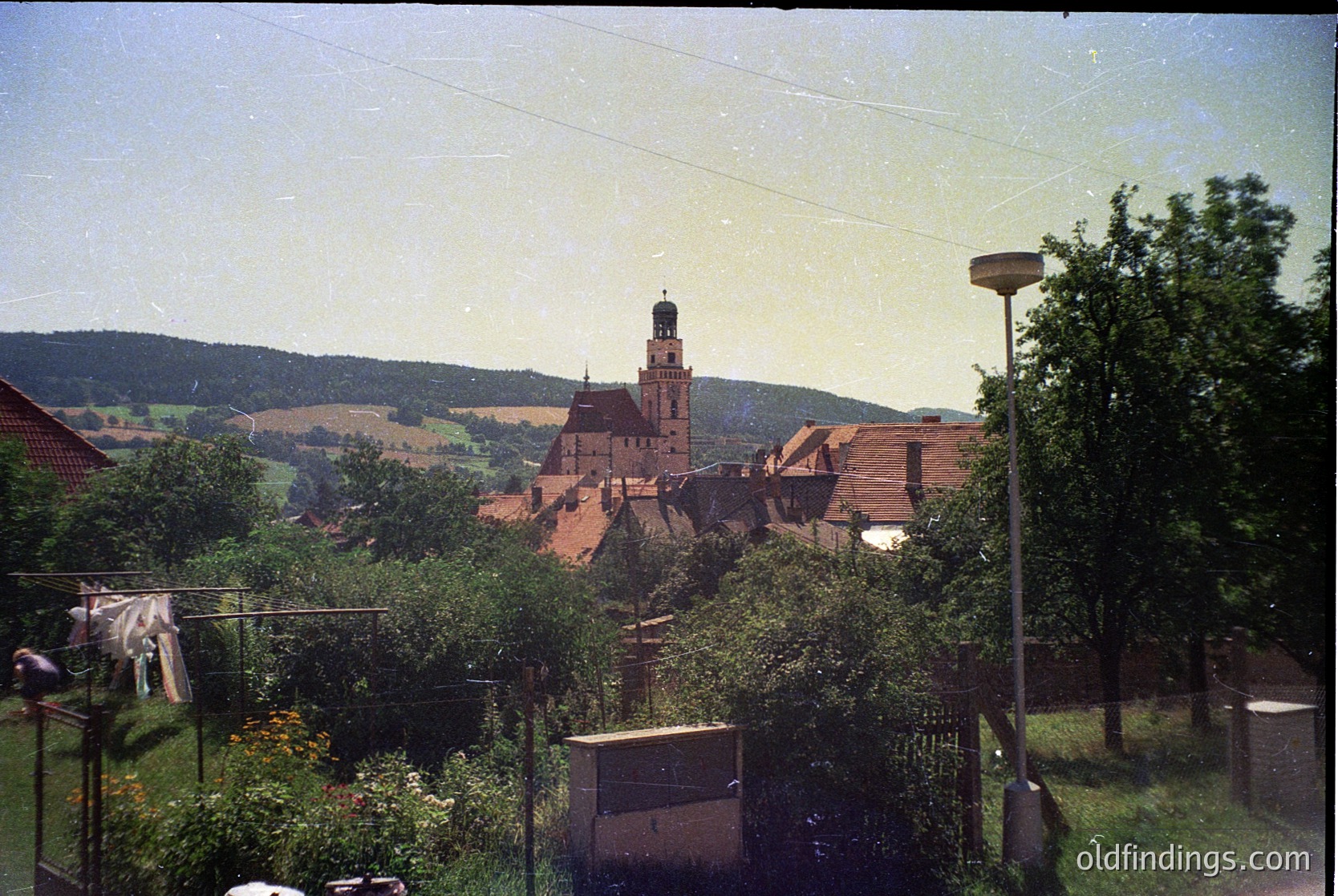 Vintage photograph of a quaint European village with a prominent red-brick church tower dominating the skyline. Foreground features lush greenery, laundry hanging on a line, and a vintage lamp post. Rolling hills and scattered houses frame the scene, suggesting rural charm. Likely late 20th century.
