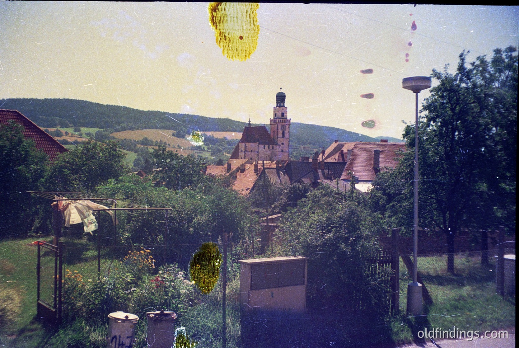 Vintage aerial view of a quaint European village with a prominent castle-like church atop a hill. Lush greenery, rustic homes, and a clothesline with garments in foreground. Color-tinted photo suggests mid-20th century (1950s–1970s).