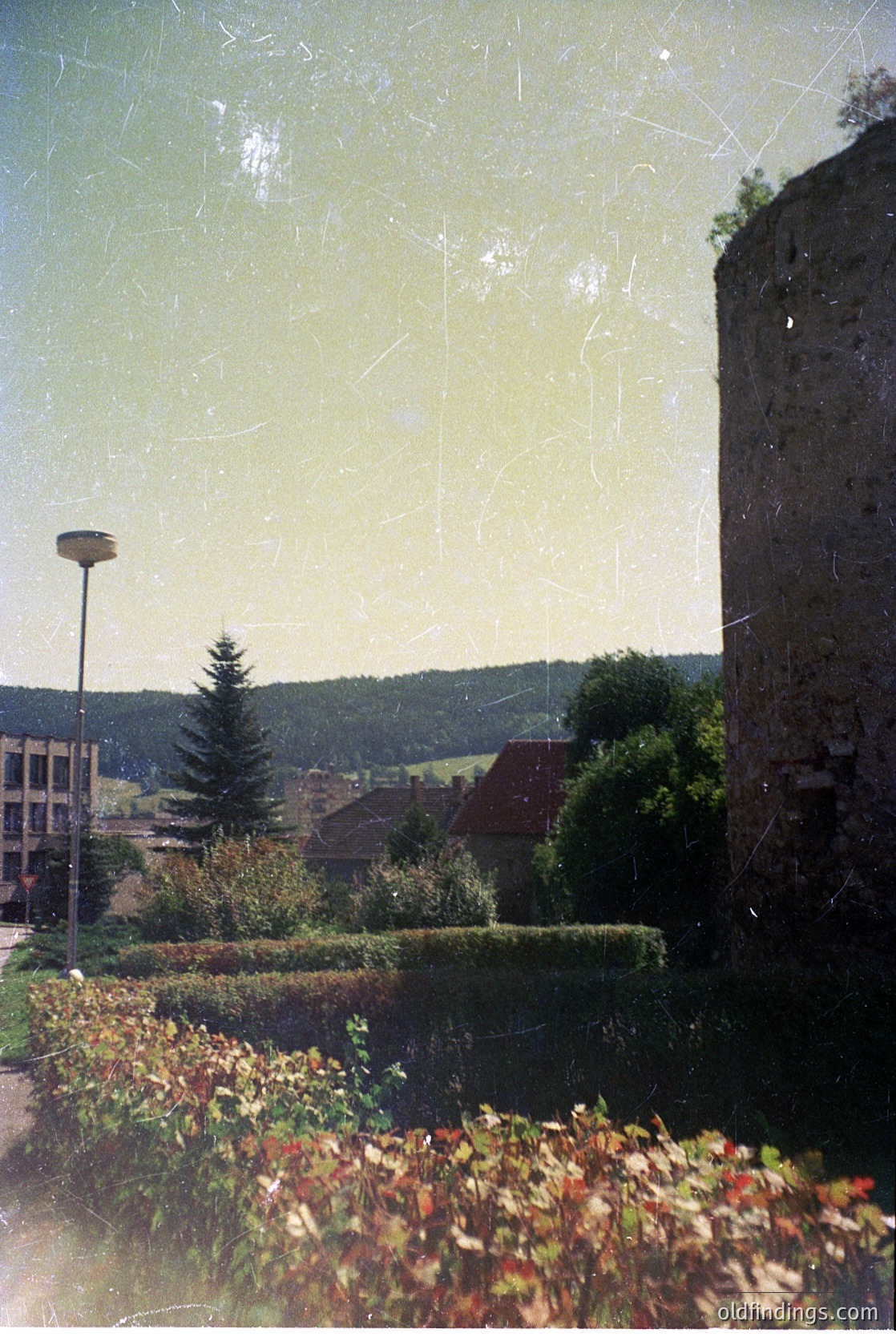 Vintage photo of a rustic European village framed by autumn foliage and a weathered stone tower. Mid-20th century architecture with sloped roofs and modern streetlight. Scenic hills in background suggest alpine or forested region.