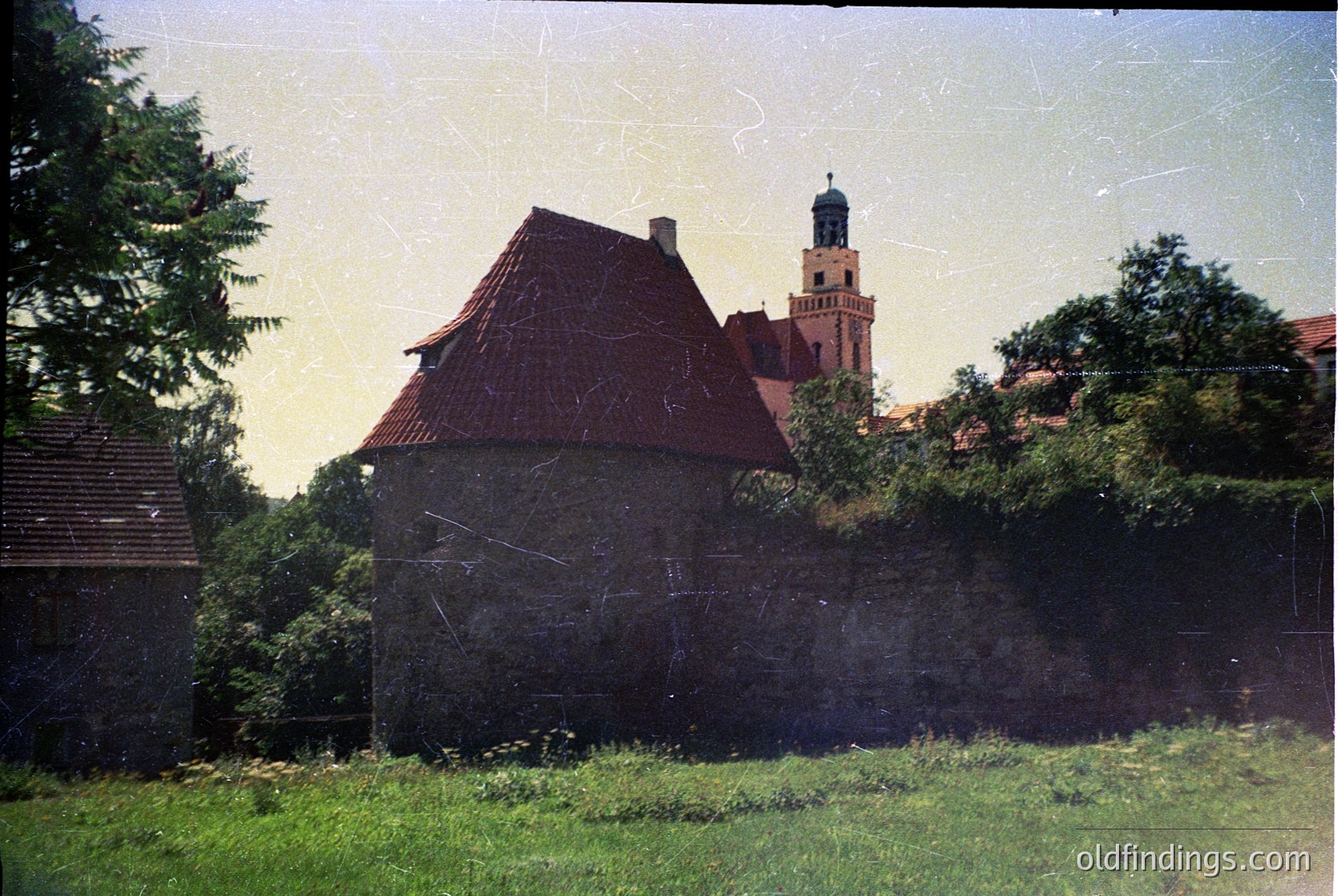 Vintage photograph of a weathered stone building with a steep red-tiled roof, surrounded by overgrown greenery. In the background, a tall church tower with a spire rises above nearby structures. Likely Eastern European rural architecture, mid-20th century.