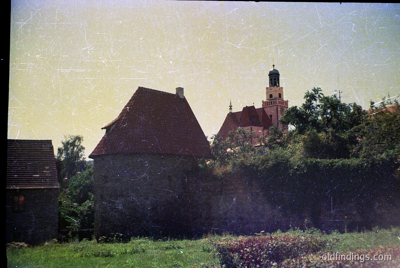 Vintage photo of a rustic stone cottage with a steep red-tiled roof, surrounded by lush greenery and a low hedge. In the background, a historic brick church with a tall spire and arched windows. Likely Eastern European countryside, mid-20th century.