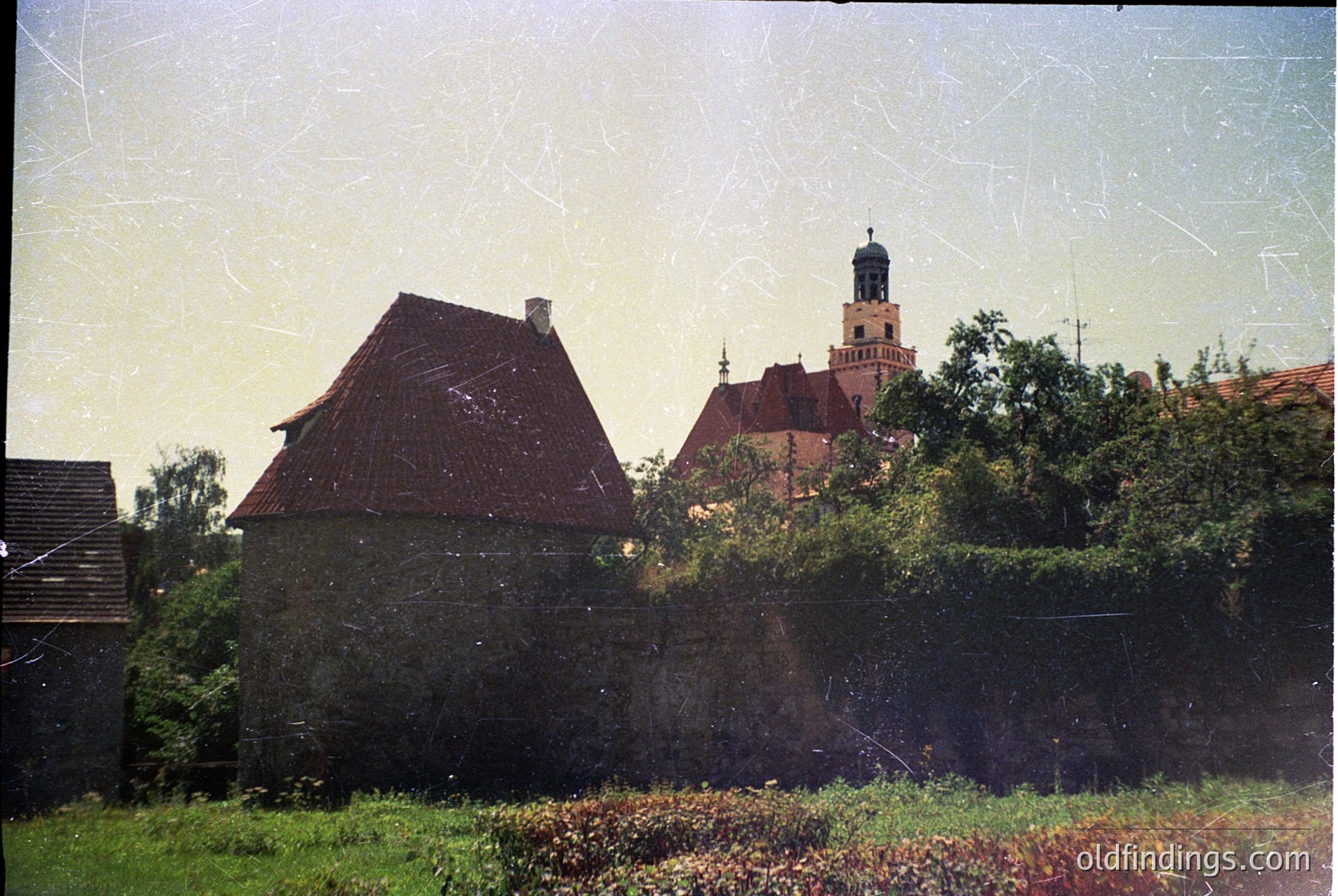 Vintage photograph of a European countryside scene featuring a rustic stone building with a steep red-tiled roof and chimney, surrounded by overgrown vegetation. In the background, a prominent church with a tall spire and green dome rises above dense foliage. The image suggests a mid-20th century rural setting, likely Eastern Europe.