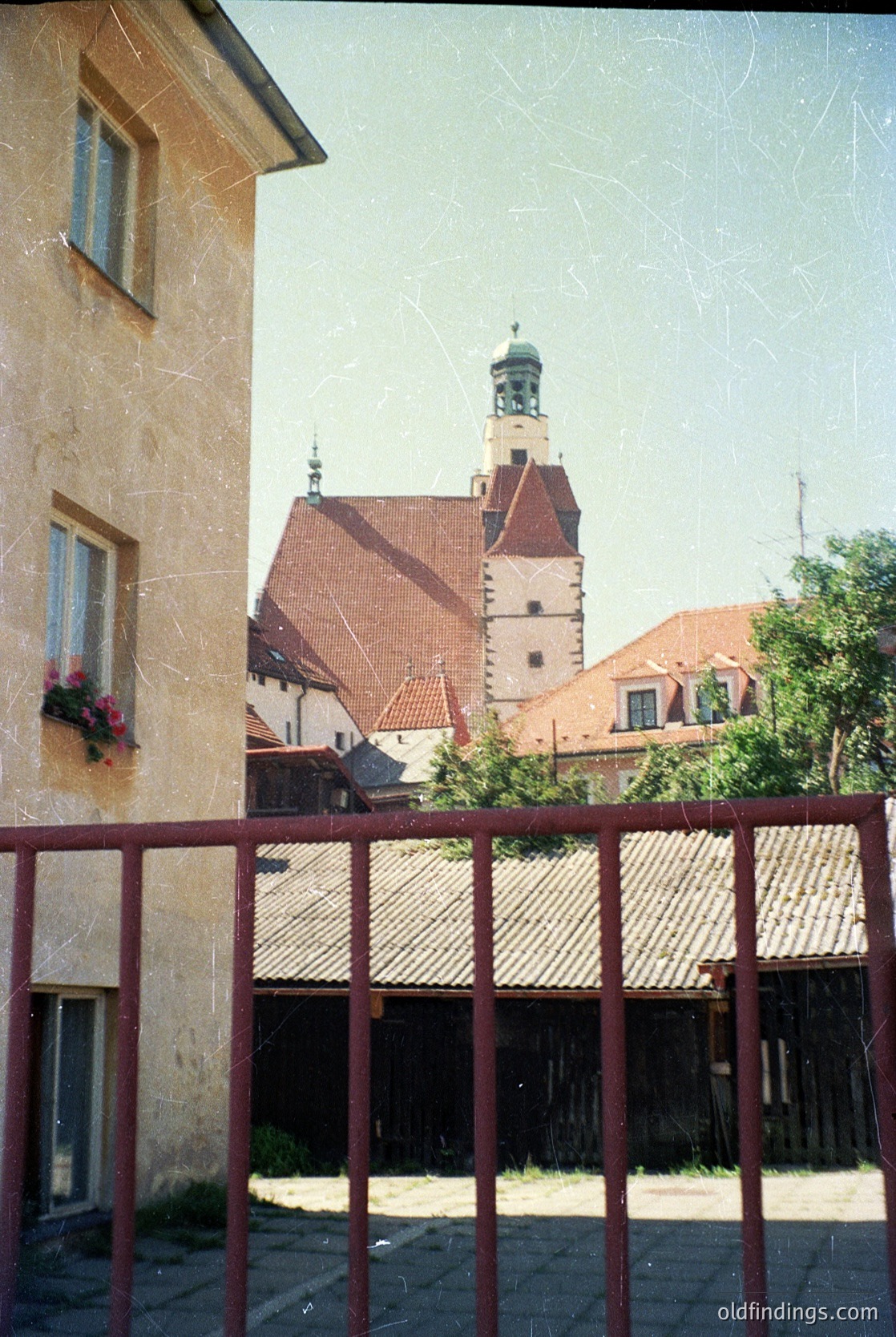 Historic European courtyard framed by rusted metal railings, showcasing a mix of stone and brick architecture. Prominent red-tiled church tower with a spire dominates the background, surrounded by smaller residential buildings. Floral window box adds color contrast. Likely 19th–early 20th century European town.