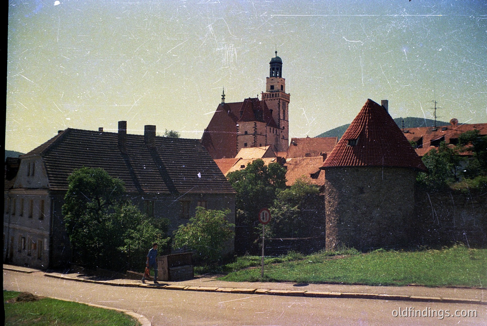 Vintage photograph of a European hilltop fortress with red-tiled roofs and a central tower, surrounded by stone walls. Lush greenery and a single pedestrian in the foreground. Likely 1960s–1980s, Eastern Bloc region.