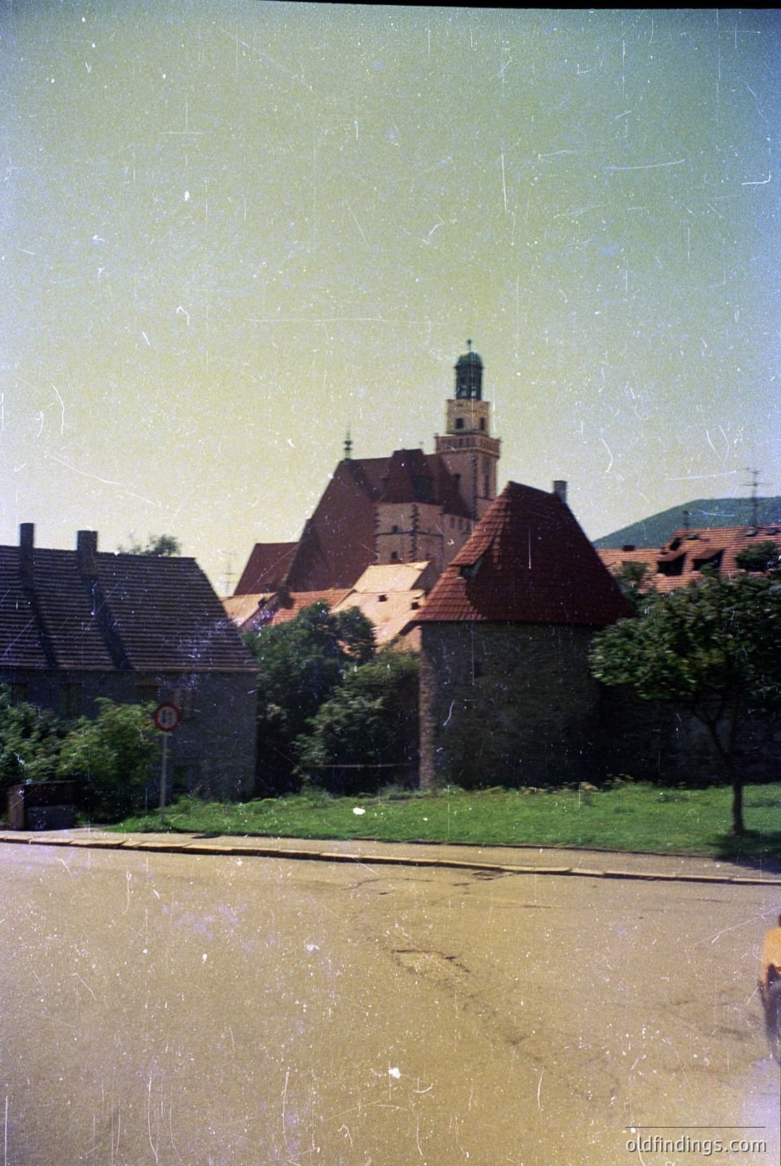 Vintage black-and-white photo of a European church with a steep gable roof and central tower, surrounded by low-pitched residential roofs. The scene suggests a small town or village setting with minimal modern infrastructure. The photo’s grainy texture and faded tones indicate it’s likely from the mid-20th century. [Historic European church with steep gable roof and central tower, mid-20th century architecture ]