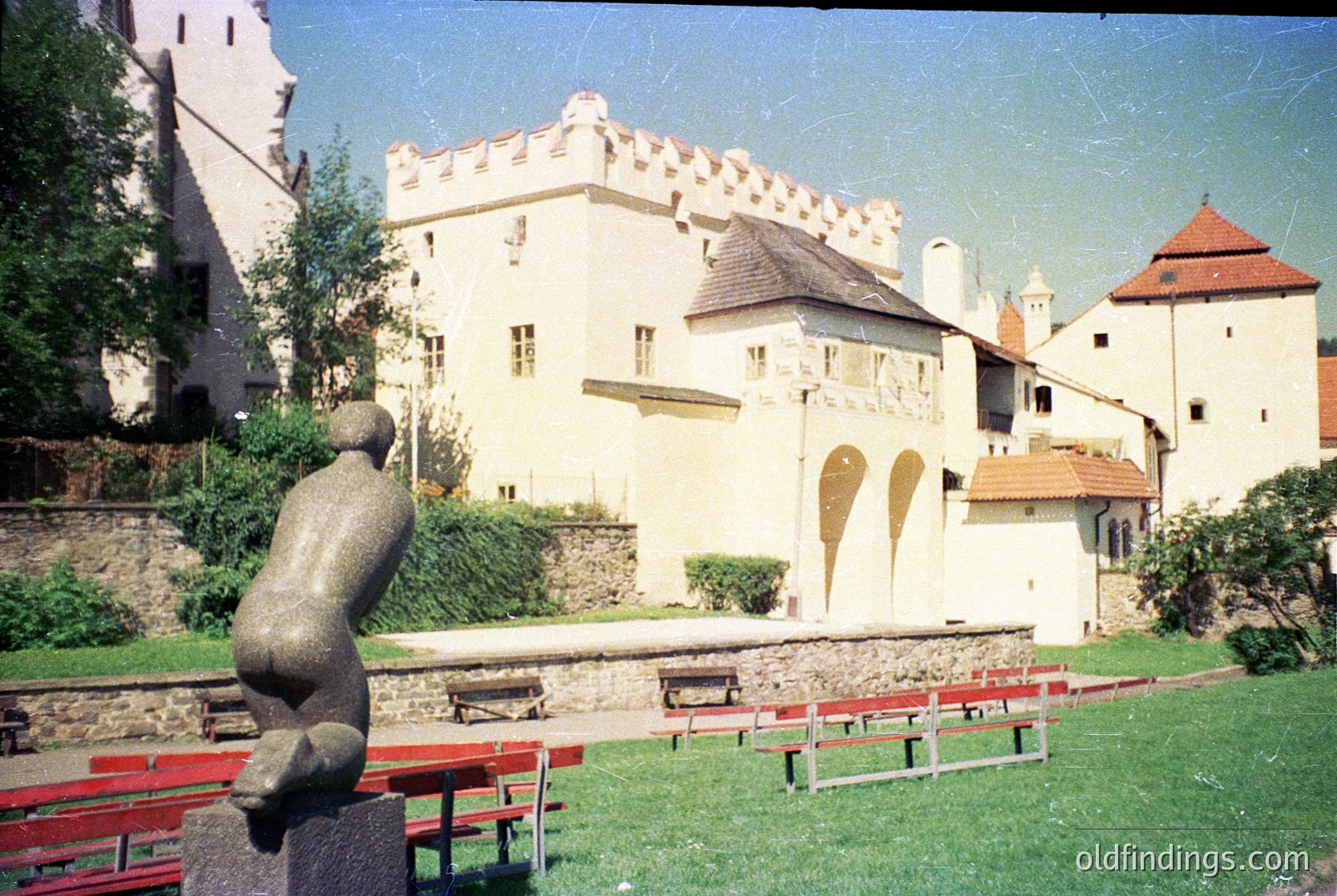 Medieval fortress with crenellated walls and red-tiled roofs, likely a European castle. Prominent stone sculpture resembling a reclining figure in foreground. Red picnic benches arranged in a courtyard with manicured lawn. Architectural style suggests 12th–16th century. Potential location: Central/Eastern Europe. [Historic European castle courtyard with medieval architecture and stone sculpture ]