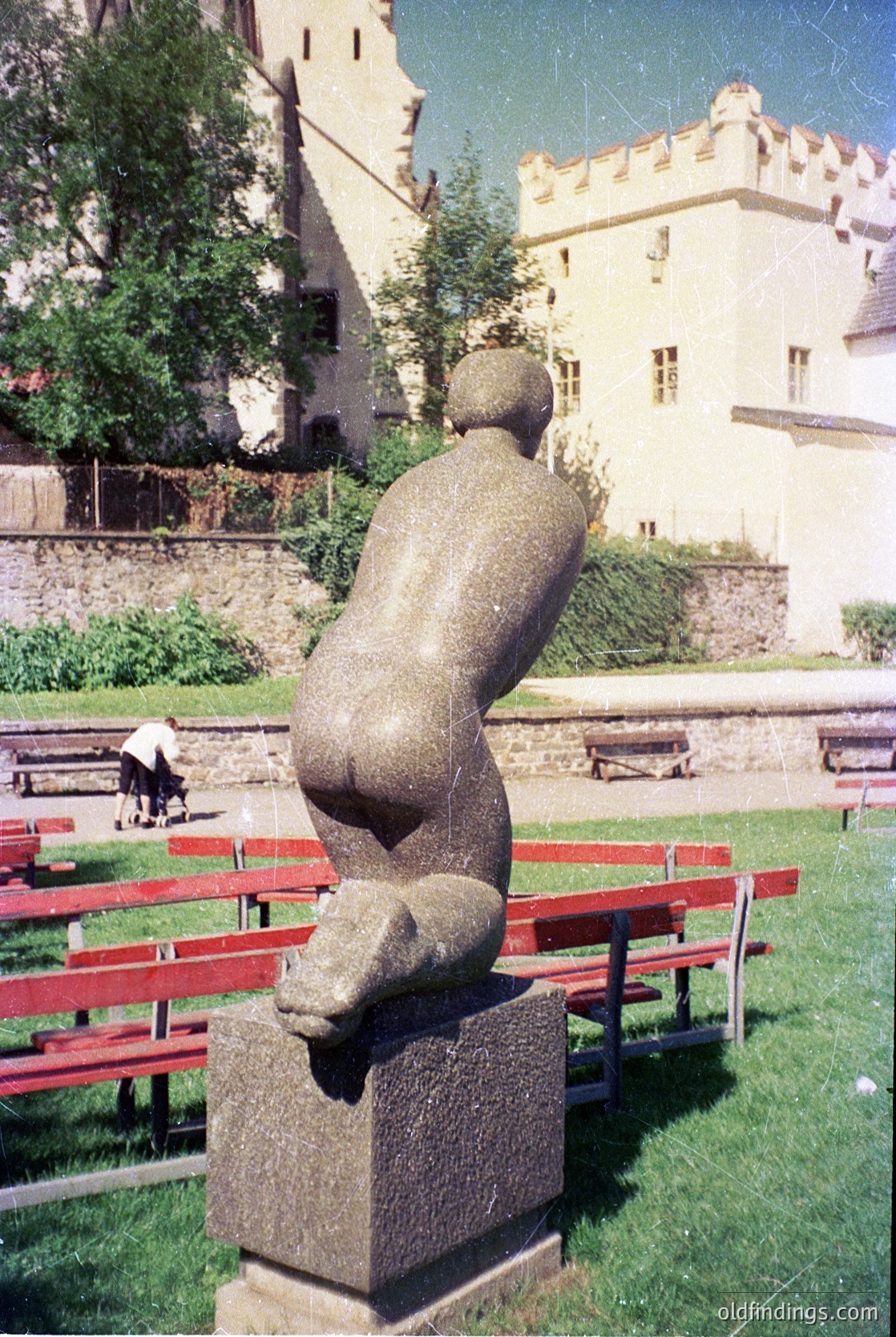 Modernist stone sculpture of seated figure in dynamic pose, placed on a square pedestal in an outdoor park setting. Red wooden benches and historic castle-style building with crenellations in background. Likely European, mid-20th century.