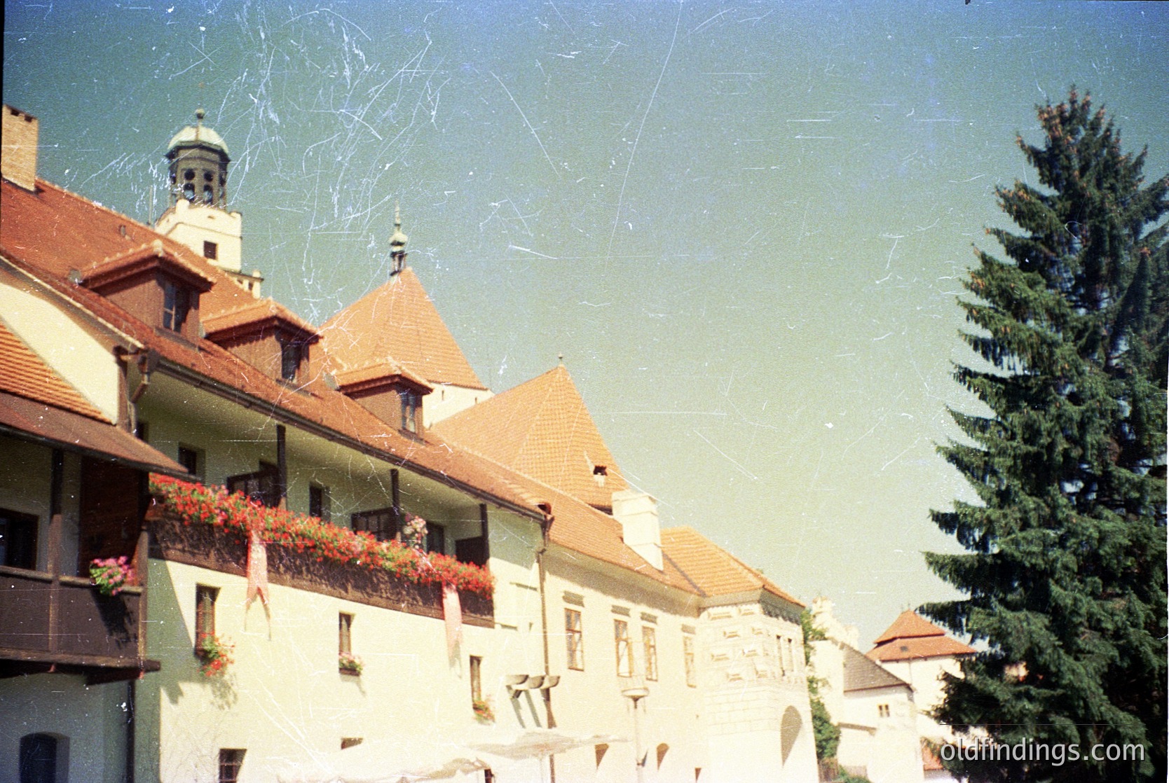 Vintage European street scene featuring a row of pastel-colored buildings with gabled roofs and flower boxes. A church tower with a dome and cross rises in the background. Signs of aging film grain and slight blur enhance nostalgic charm. Likely or European architecture.