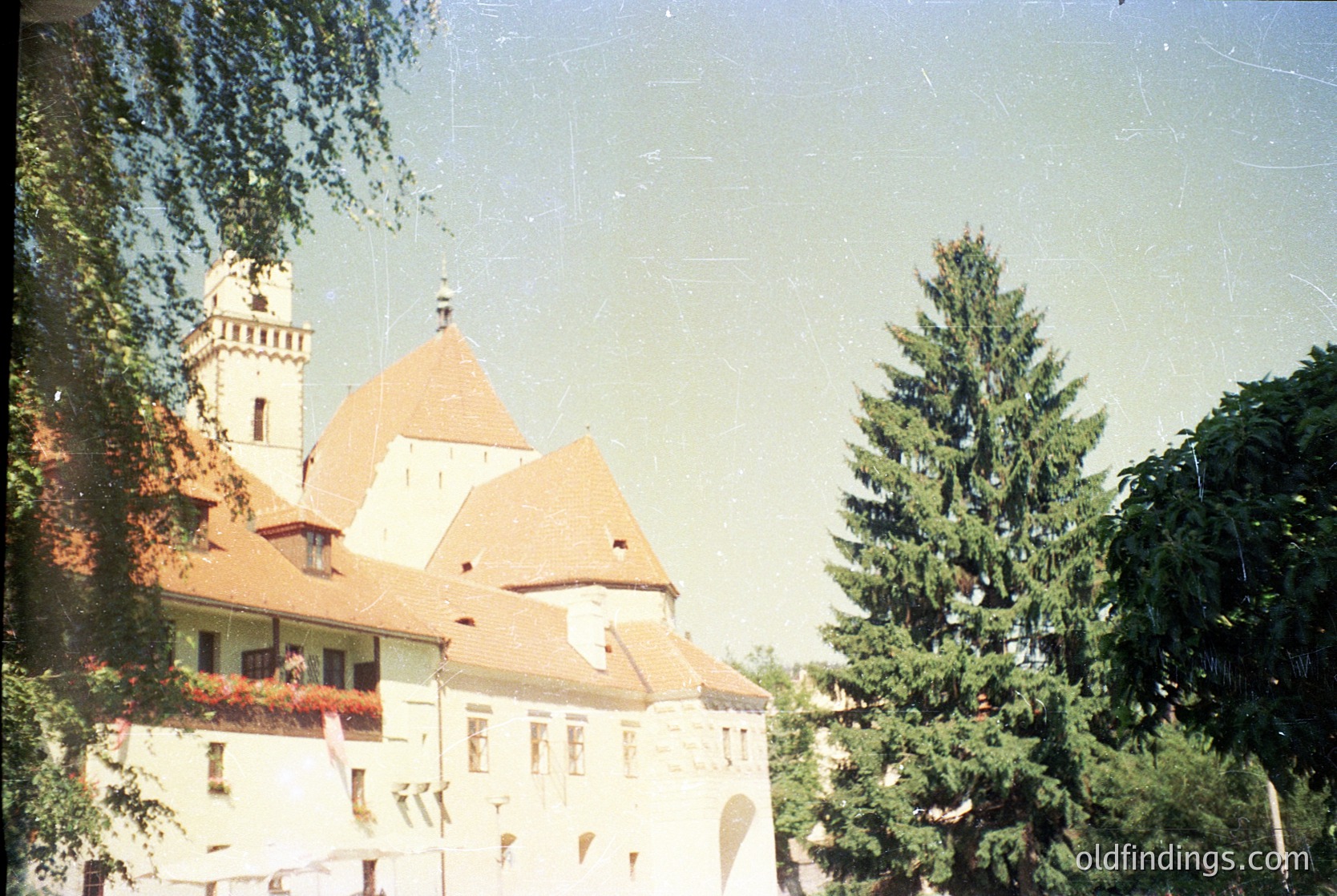 Vintage photograph of a European castle-style building with a conical roof and tower, surrounded by lush greenery. The structure features arched windows and decorative flower boxes. Likely late 20th century, possibly . The aged film grain and sepia tone suggest archival or nostalgic value.