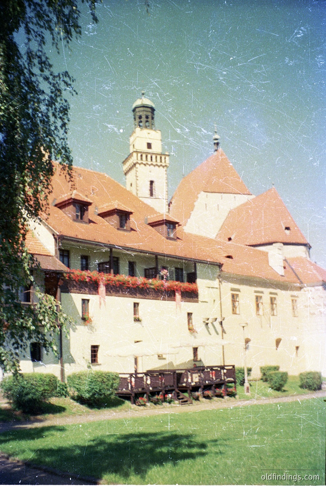 Historic European castle with red-tiled gables and central tower, featuring floral balconies. Likely 19th-century architecture with well-manicured grounds and vintage wooden cart.