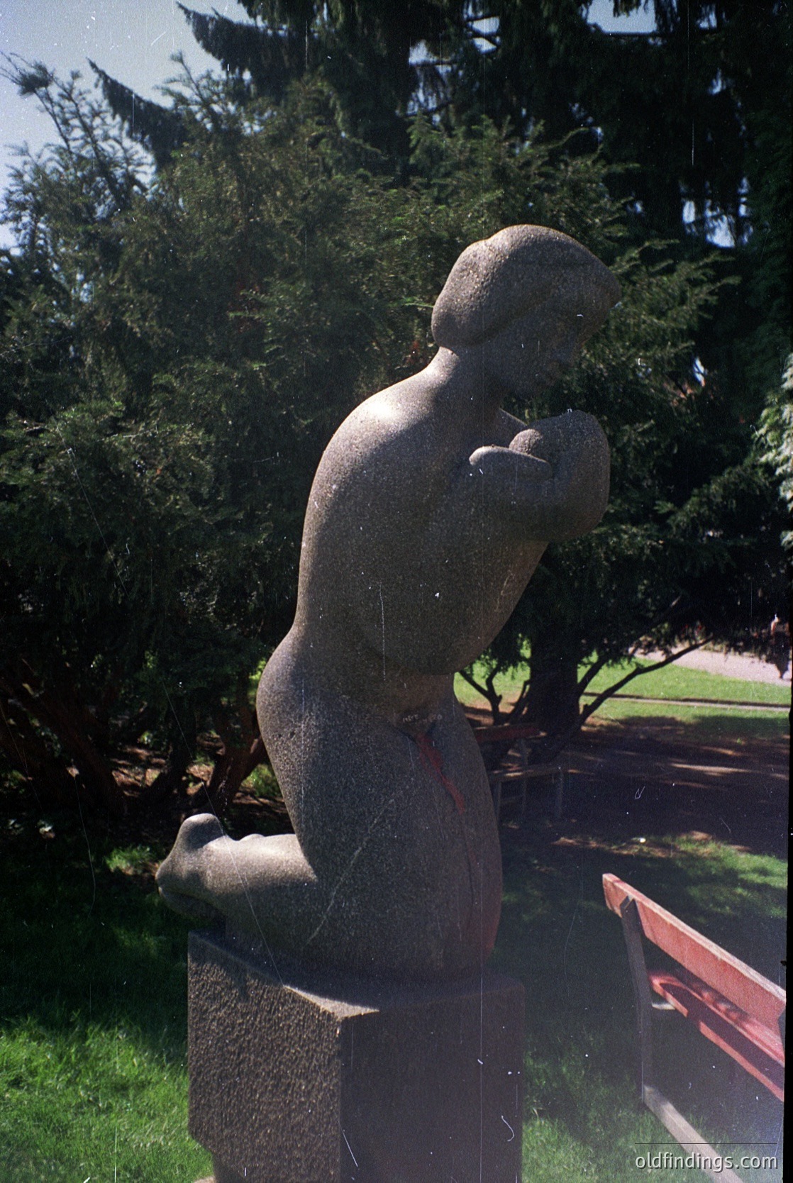 Bronze abstract sculpture of a seated figure with exaggerated, rounded forms, set on a textured pedestal in a landscaped park. The stylized pose suggests mid-20th century modernist influence.