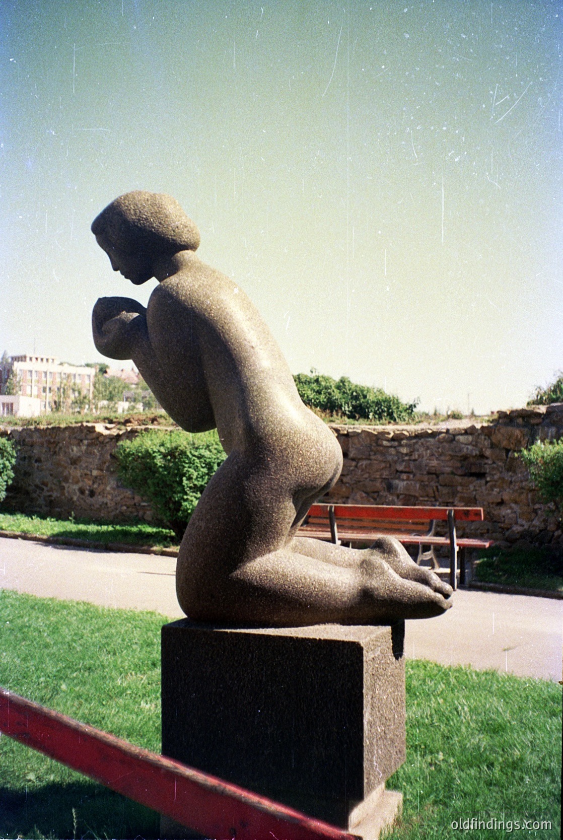 Abstract seated stone sculpture in outdoor park setting, likely . Minimalist human form with head tilted forward, hands clasped. Positioned on concrete pedestal with red metal railing in background. Urban greenery and stone wall suggest public space. -1980s