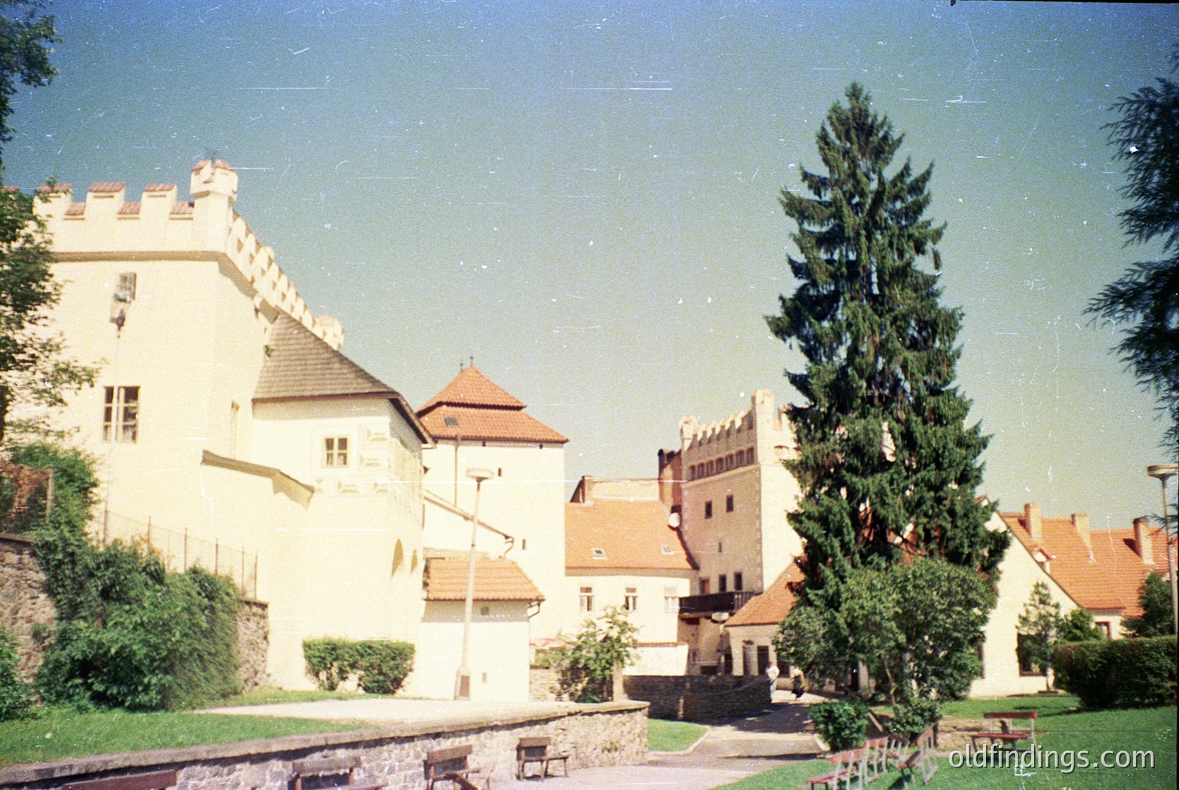 Medieval-style castle complex with crenellated towers, red-tiled roofs, and stone walls. Central courtyard features a tall evergreen tree and a small fountain. Likely European, possibly architecture. Ideal for historical research or fantasy-themed design references.