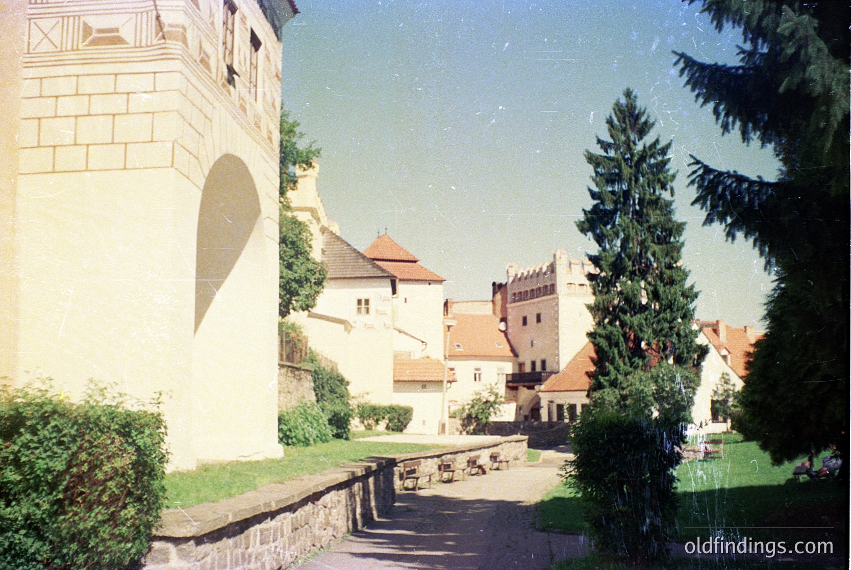 Medieval-style courtyard with stone tower and red-tiled roofs, framed by evergreen trees. Likely European castle or historic estate, 19th–early 20th century. Stone benches and paved pathways suggest public or tourist access.