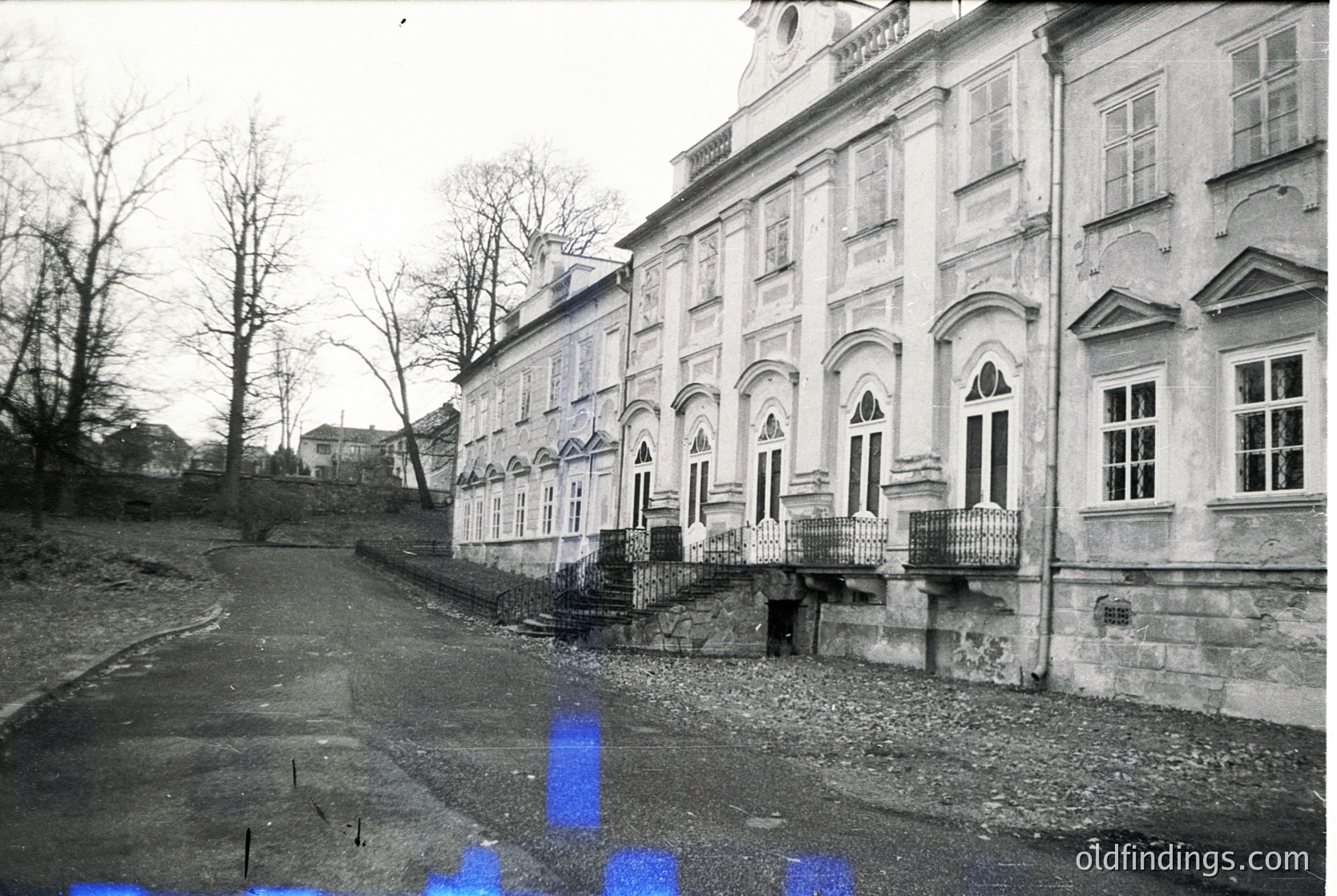 Neoclassical building with arched windows and decorative cornices, likely from the early-to-mid 20th century. Symmetrical façade features wrought-iron balcony railings and a central entrance. Gravel driveway curves toward the structure, bordered by sparse trees. Potential institutional or public use.