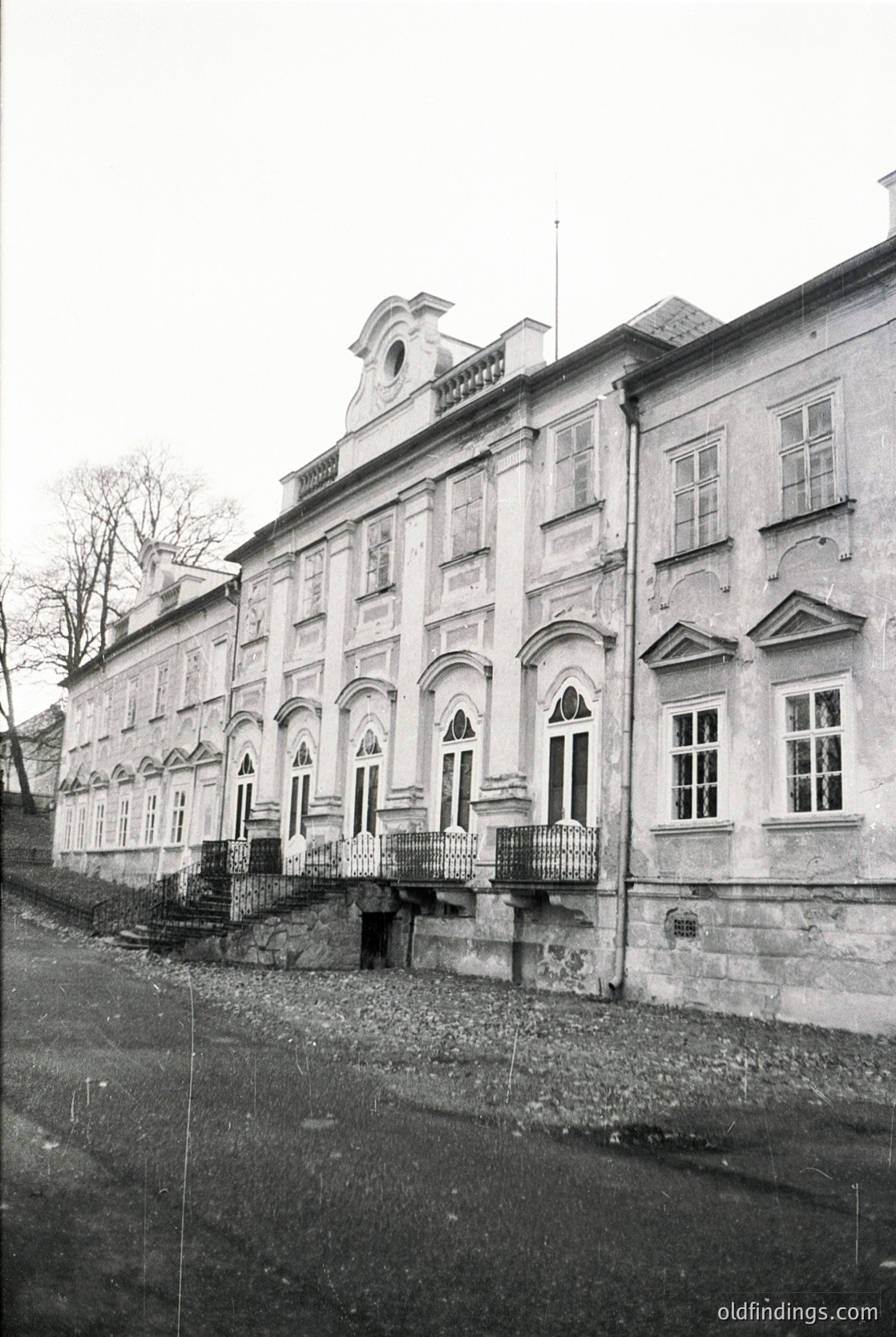 Neoclassical stone building with arched windows and decorative cornices, likely a historic institutional or public structure. Symmetrical façade features a central entrance with a balcony above. Cobblestone street and minimalist urban setting suggest European architectural heritage.