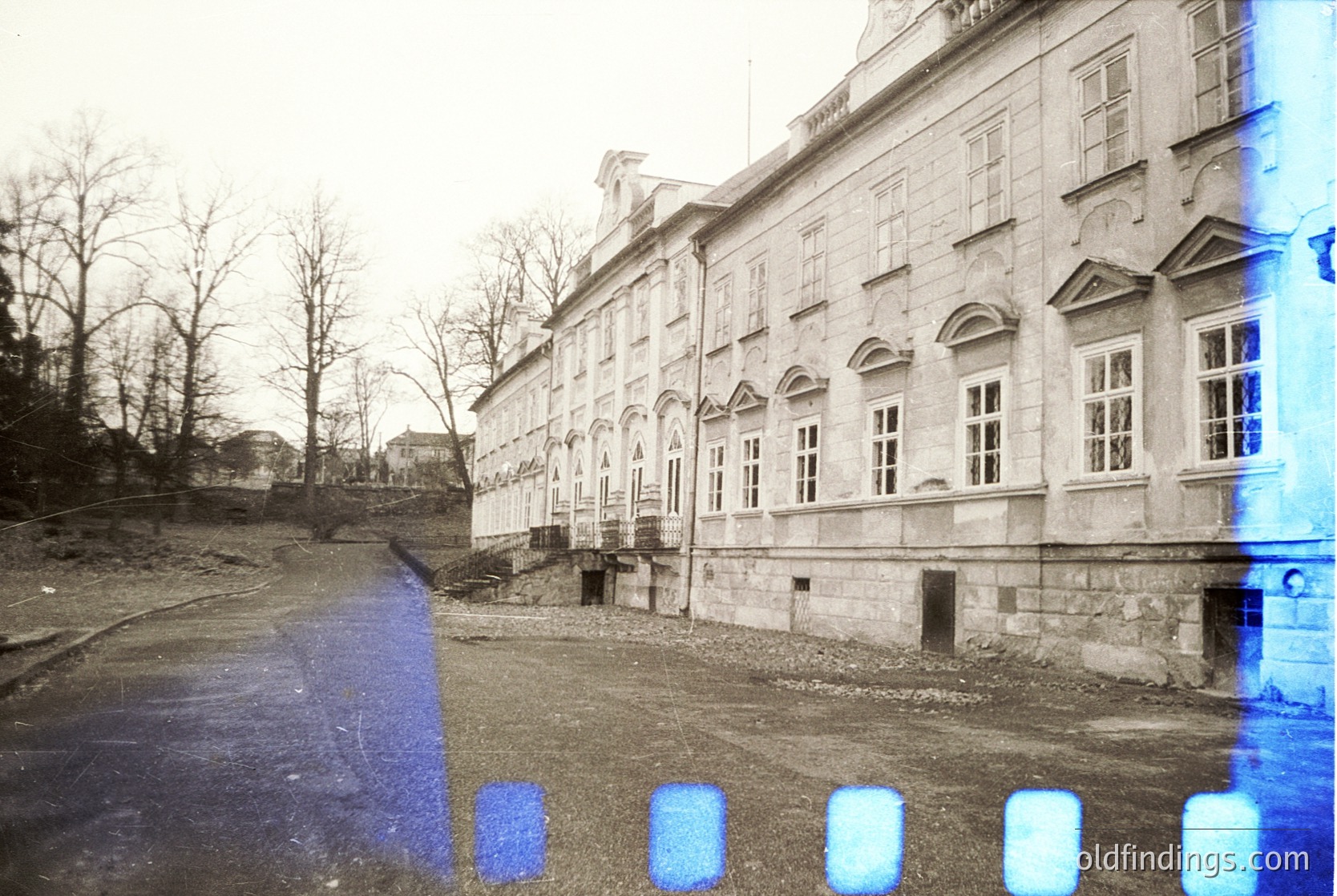 Neoclassical stone building with arched windows and decorative cornices, captured on aged film. Empty courtyard with bare trees, suggesting late autumn/winter. Overlaid blue tint and film grain indicate vintage photography, likely mid-20th century. Architectural details suggest institutional or governmental use.
