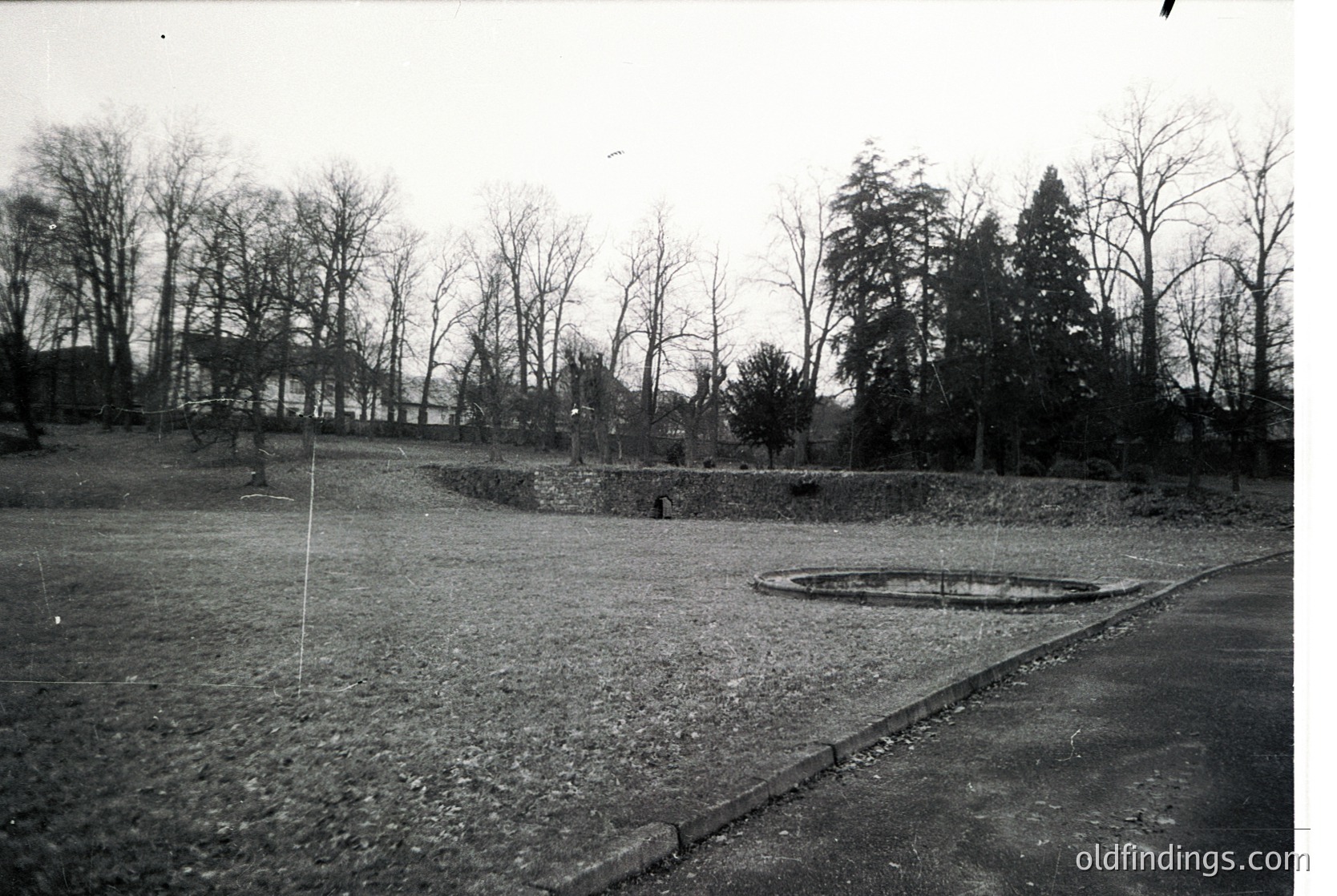 Black-and-white photograph of a barren, open courtyard surrounded by bare trees and low stone walls. Circular concrete structure in center, likely a well or monument. Asphalt path curves along right edge. Mid-century architectural style, possibly institutional or public space.