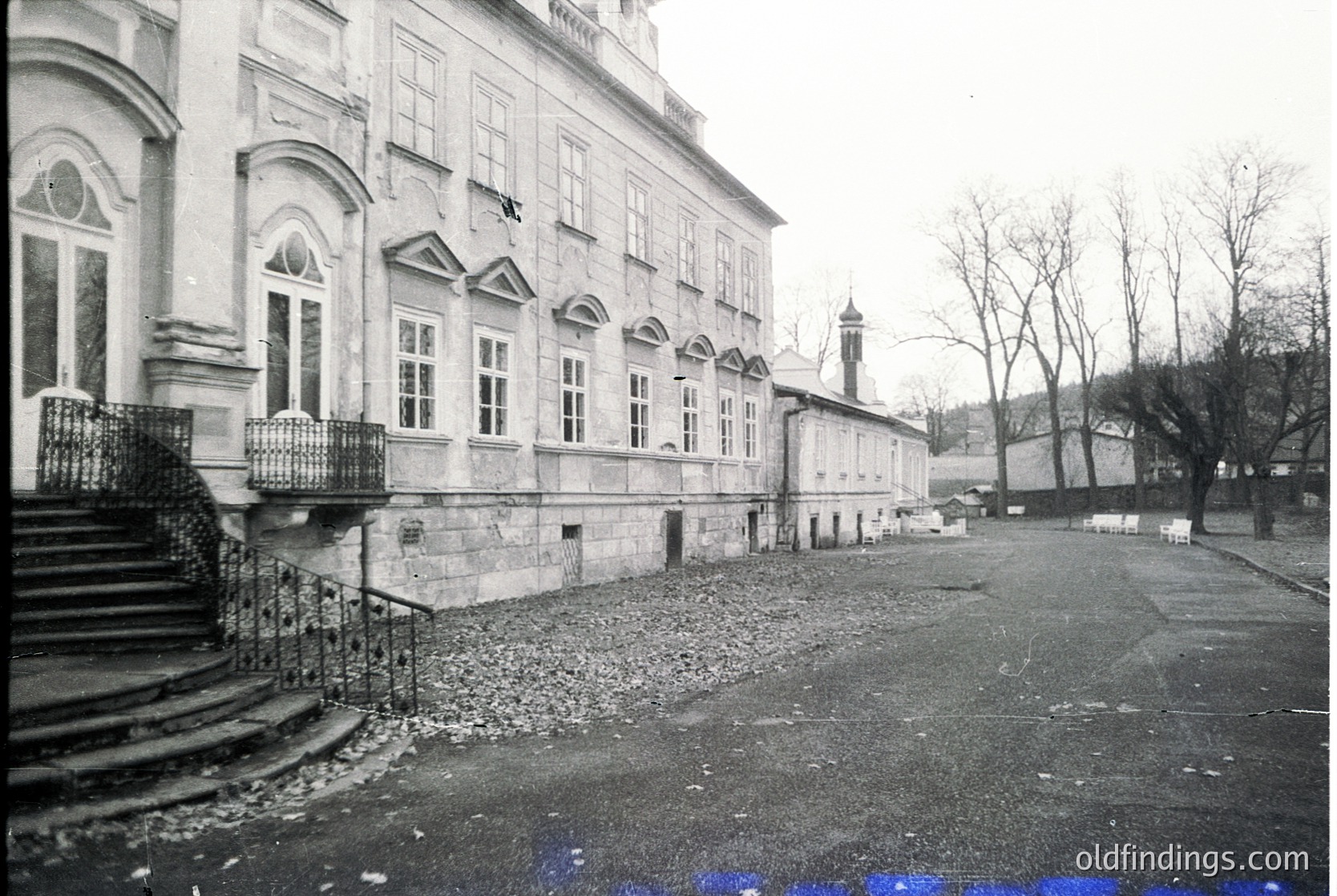 Neoclassical building façade with arched windows and wrought-iron balcony railings, likely from the early 20th century. Gravel courtyard and stone steps lead to an entrance. Minimalist, symmetrical architecture with a distant tower and bare trees.