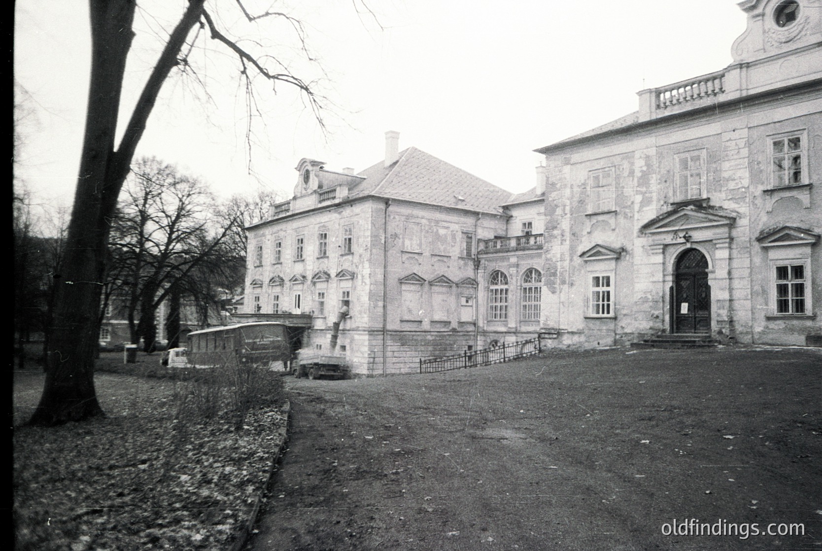 Neoclassical stone building with symmetrical façade, arched doorway, and rectangular windows. Courtyard entrance flanked by balustrades. Bare trees and gravel ground suggest late autumn/winter. Likely institutional or public use.