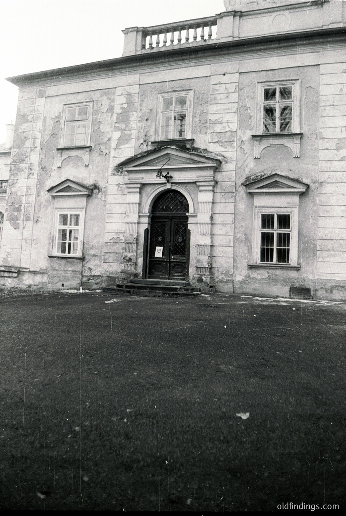 Neoclassical stone façade featuring a central arched doorway with wrought-iron gates, flanked by symmetrical rectangular windows. Weathered surfaces suggest aged construction, likely mid-20th century. Balustrade atop the roofline hints at formal European architectural influence.