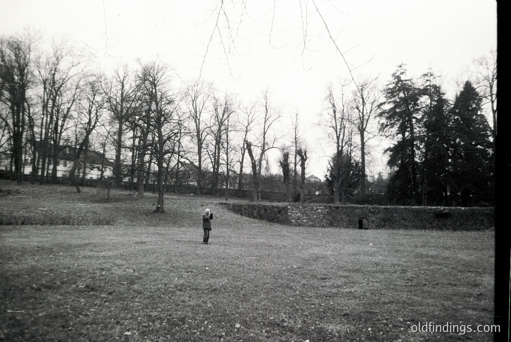 Black-and-white mid-20th century landscape featuring a lone figure standing on a grassy field bordered by a low stone wall. Leafless trees and a distant building suggest late autumn/winter. Overhead wires and a rural setting indicate possible agricultural or suburban area.