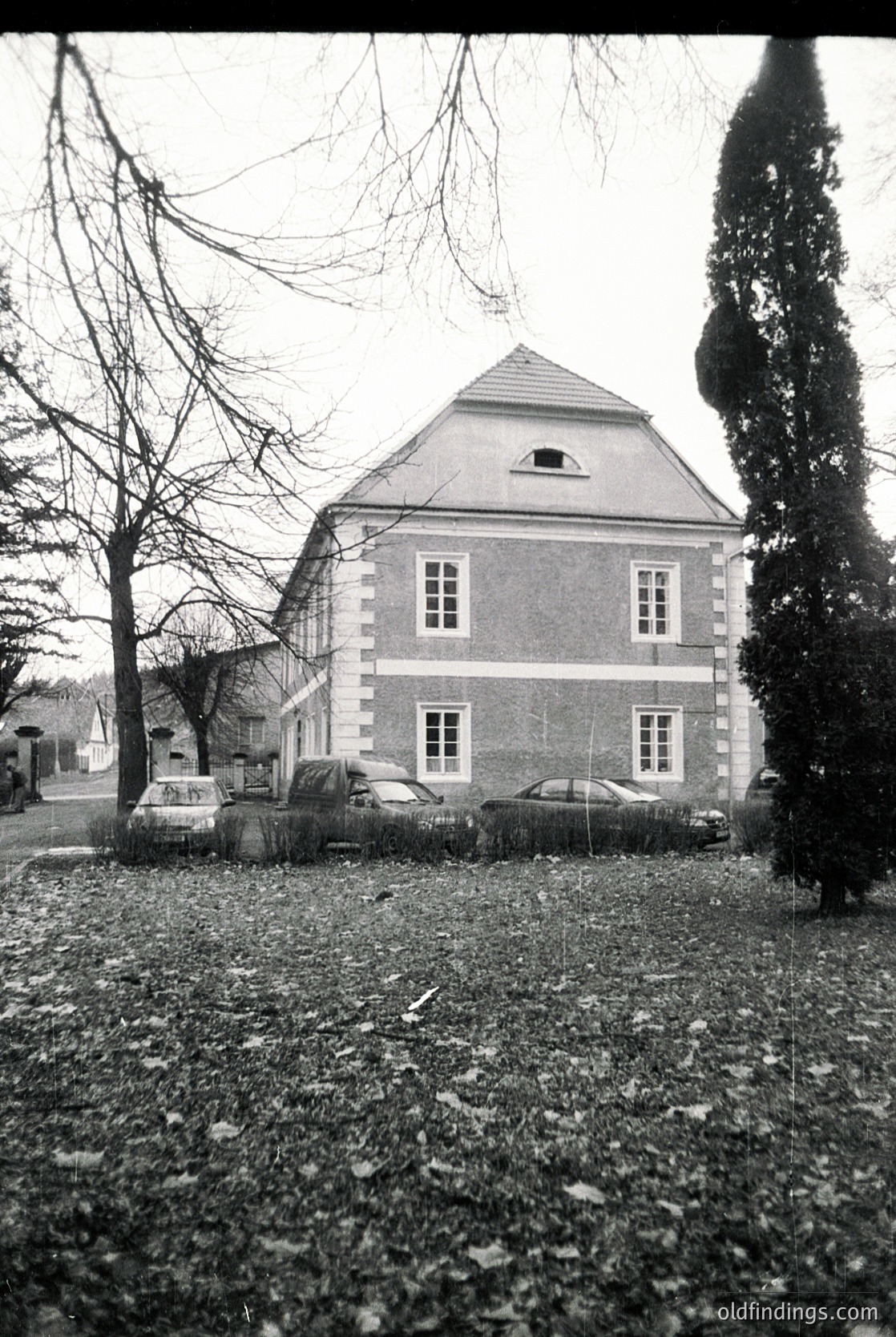 Two-story brick house with gabled roof, symmetrical window placement, and a small dormer. Classic 1950s–1960s European suburban architecture. Vintage cars parked in front, likely pre-1970s models. Leafless trees suggest late autumn/winter.