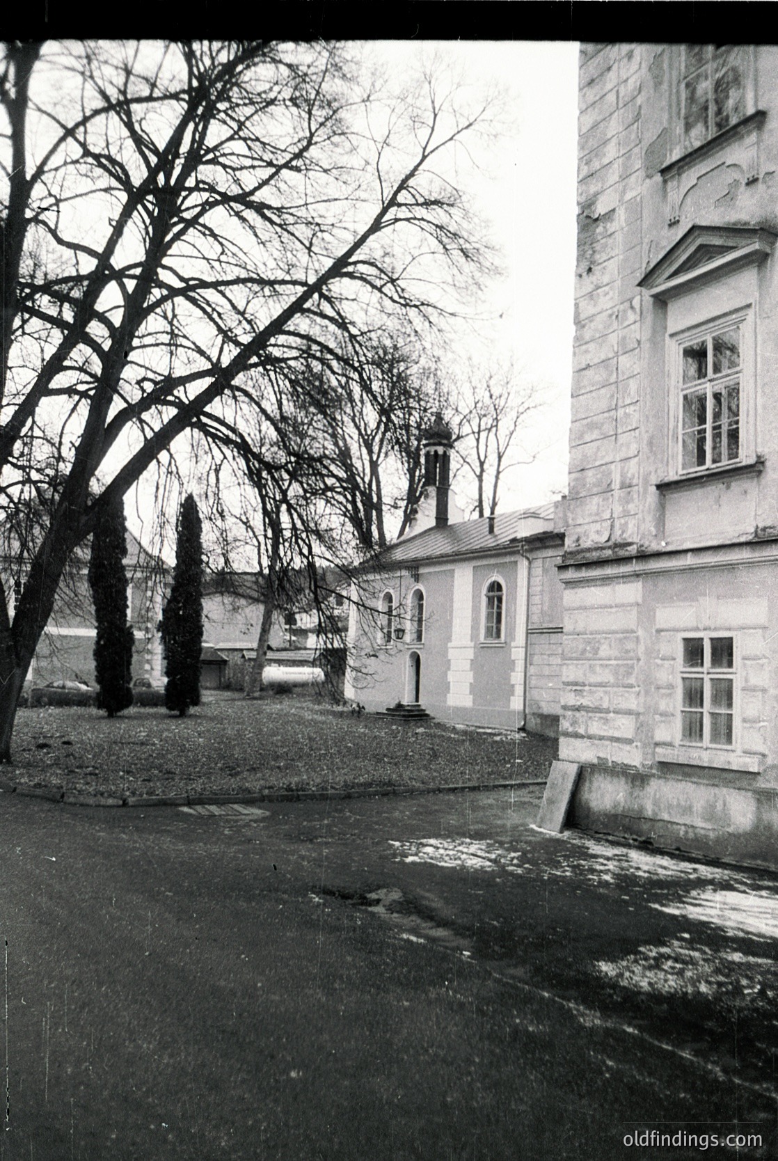 Vintage black-and-white photo of a courtyard featuring a stone church with arched windows and a bell tower, surrounded by leafless trees. The building’s weathered facade suggests mid-20th century European architecture.