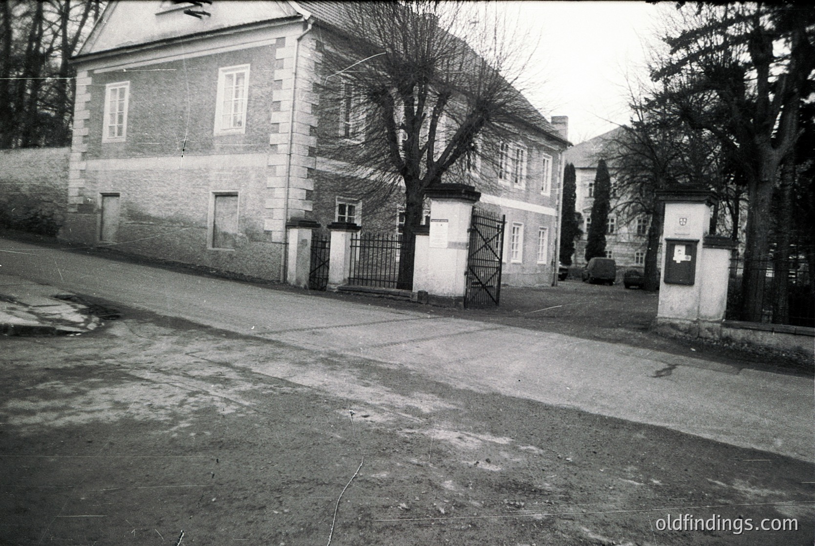 Two-story institutional building with stone façade and wrought-iron gates, likely mid-20th century. Courtyard entrance flanked by gated arches; bare trees suggest winter or early spring. Asphalt road and minimal signage indicate rural or suburban setting.