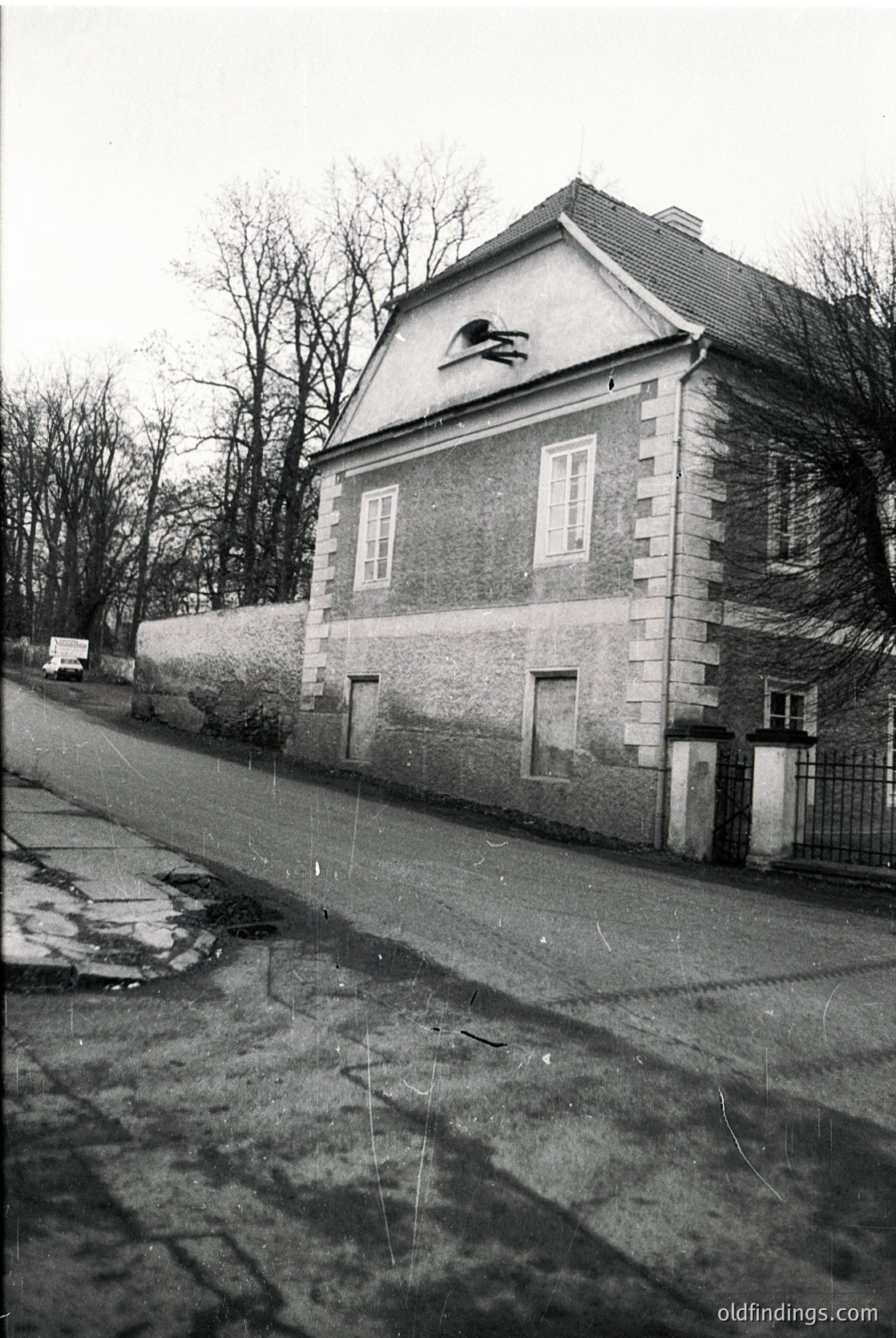 Two-story brick building with gabled roof, featuring a decorative stone cornice and a small relief sculpture near the roofline. Wet cobblestone street with puddles, bordered by a high stone wall. Leafless trees and minimalist urban setting.