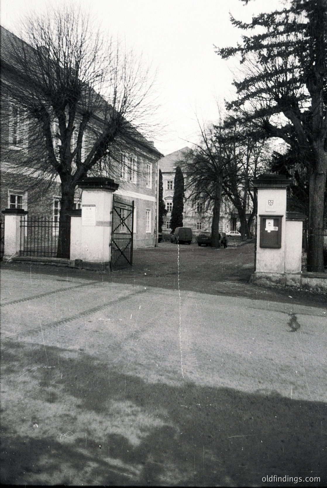 Mid-century European street scene with Soviet-era architecture. Symmetrical gatehouse with "1953" plaque and "1954" sign, flanked by leafless trees. Classic Soviet-style buildings in background, likely Eastern Bloc (1950s–1960s). Wet pavement suggests recent rain.