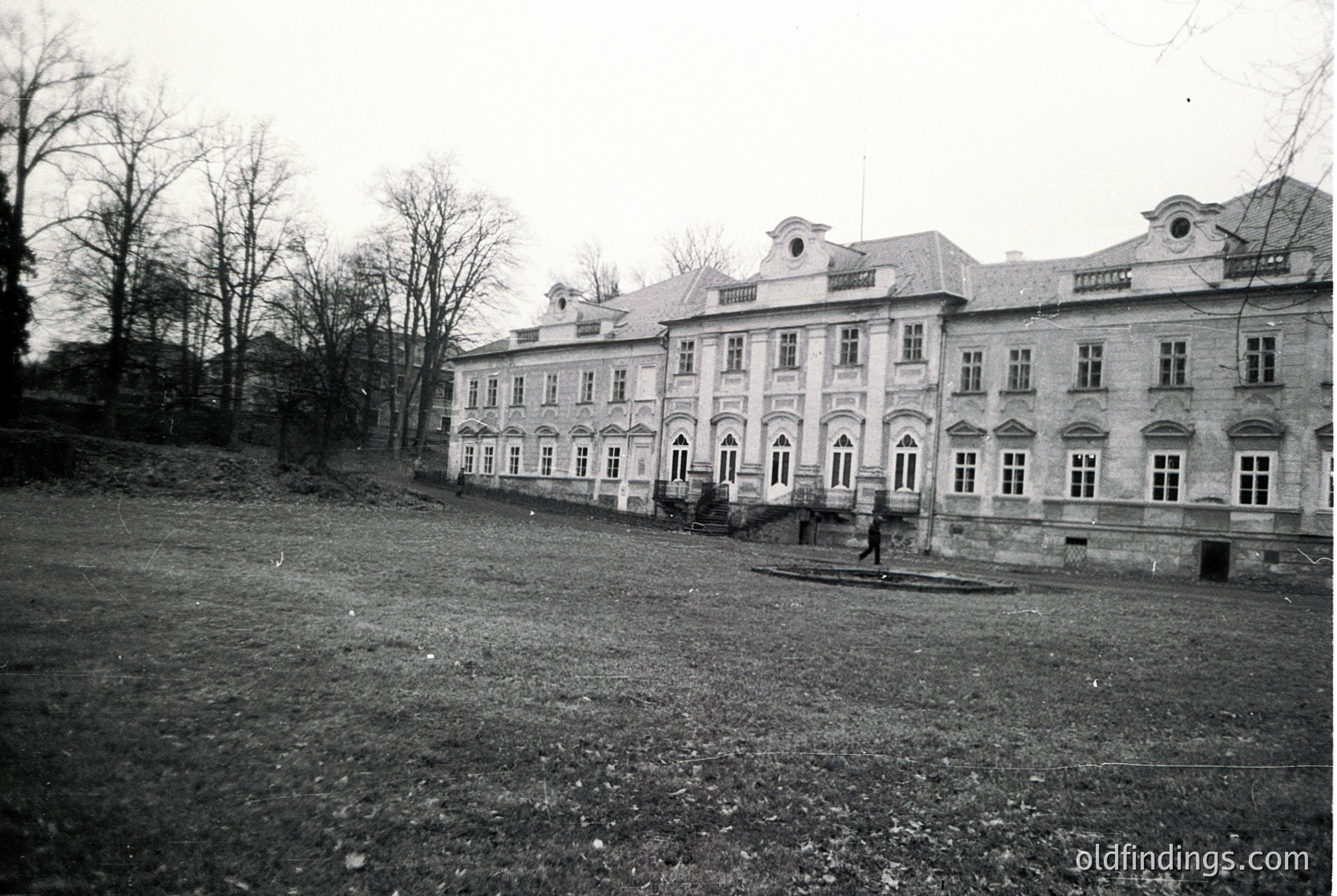Neoclassical building with symmetrical façade, arched windows, and balustrade details. Likely early-to-mid 20th century European architecture. Open courtyard with sparse vegetation and a lone figure in motion.