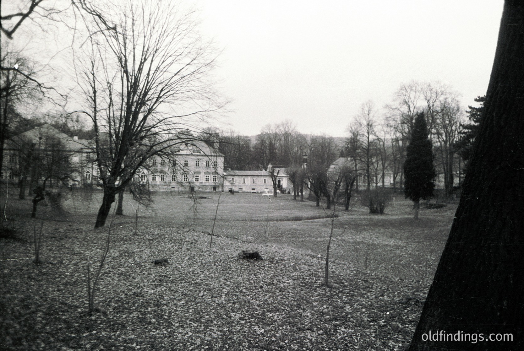 Neatly manicured lawn bordered by bare winter trees frames a two-story classical-style building with symmetrical windows and a central entrance. Overcast sky enhances muted tones in this vintage black-and-white shot.