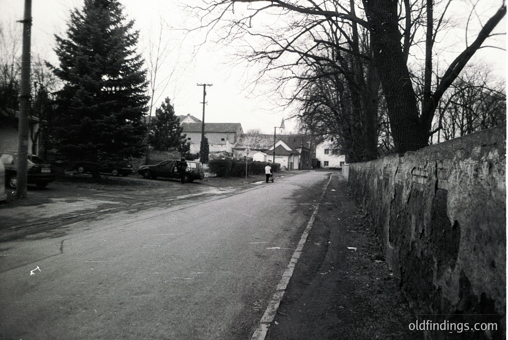 Vintage black-and-white street scene featuring a quiet residential road flanked by a stone wall and sparse trees. Mid-century architecture visible in background with a single-story house and church steeple. Classic 1950s–1960s street lamp and parked vintage car on left.