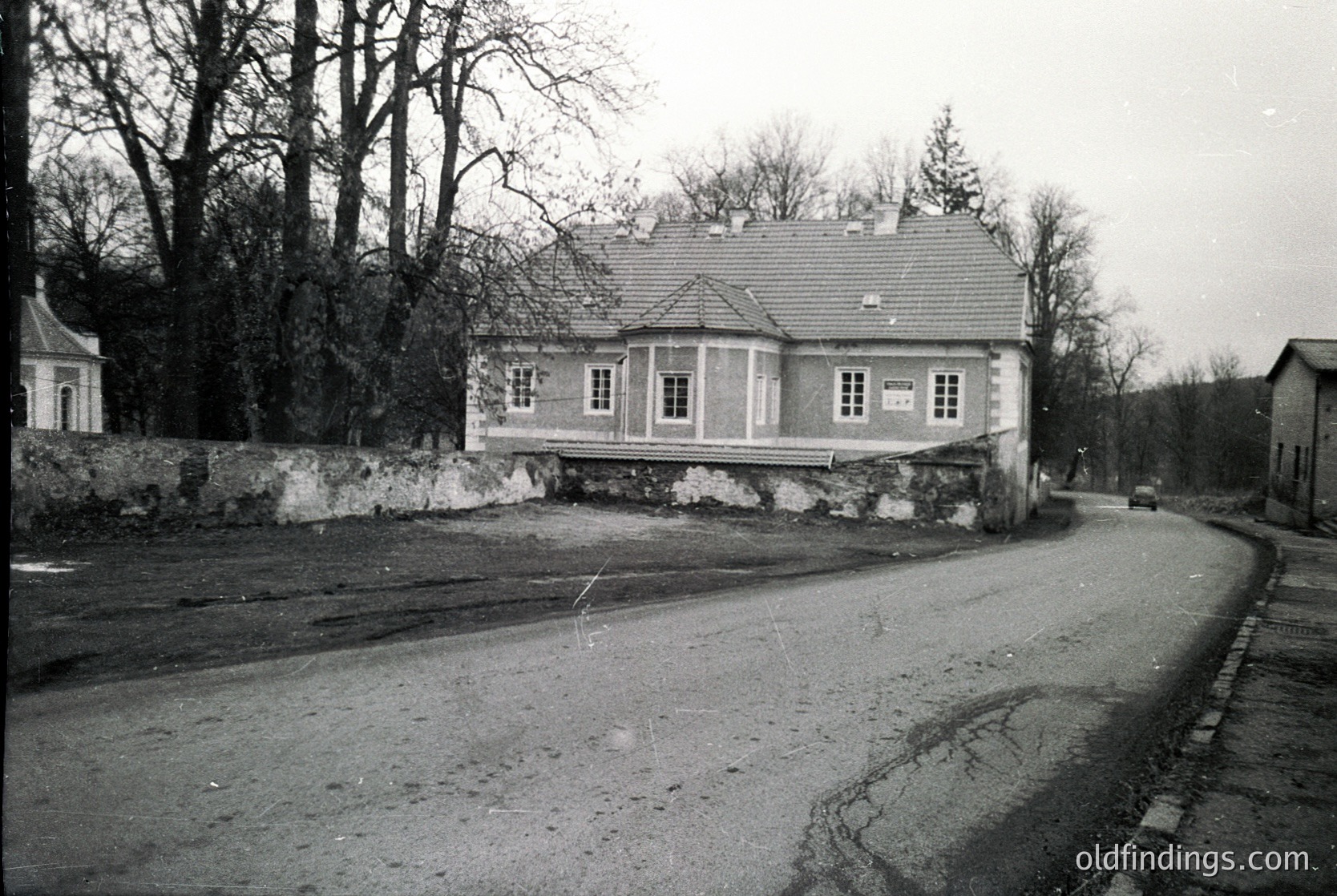 Two-story villa-style building with symmetrical windows, set on a stone foundation. Asphalt road curves left with cobblestone sidewalk. Leafless trees suggest late autumn/winter. Black-and-white, likely mid-20th century.