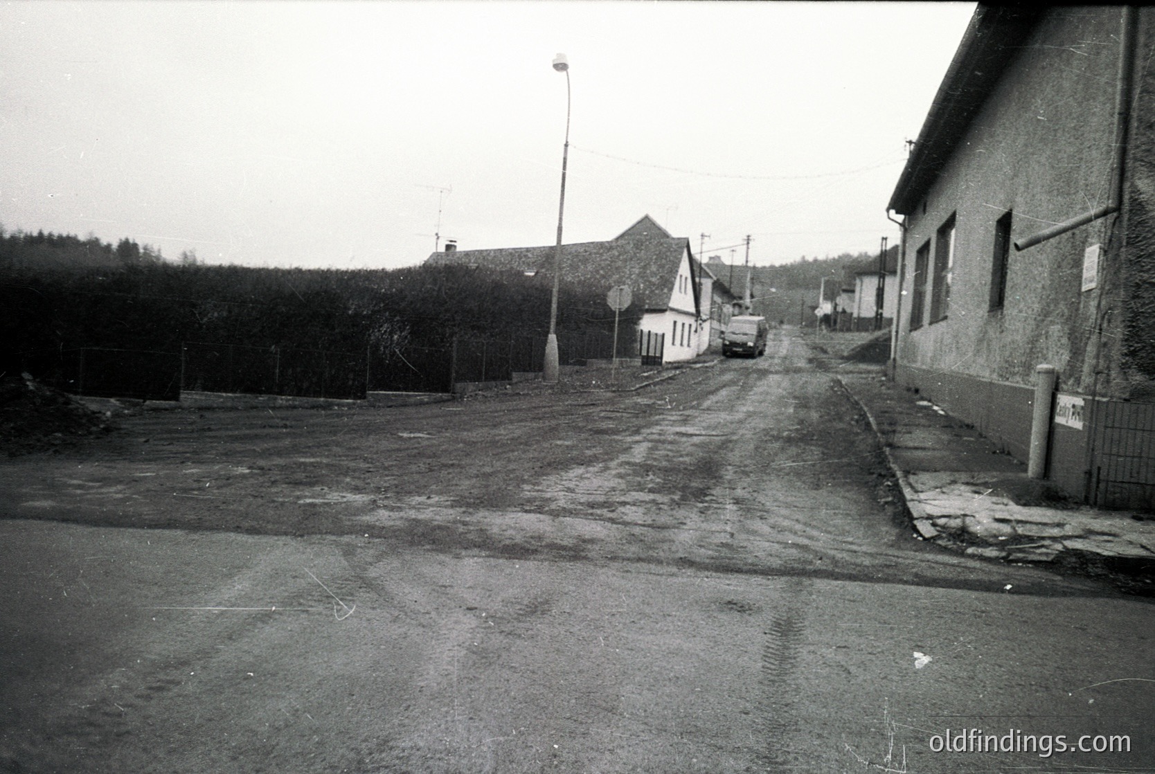 Empty rural street with unpaved sidewalks, lined by utilitarian buildings and sparse vegetation. Mid-century concrete road surface with visible cracks. Single-story houses and a small white structure in background. Overcast sky suggests early 20th century or mid-century European village.
