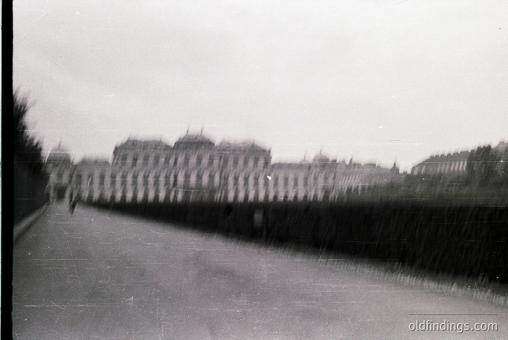 Blurred monochrome street scene with indistinct figures walking alongside a high, dense hedge. Distant buildings suggest urban architecture, likely European. Motion blur indicates handheld or slow shutter speed. Potential mid-20th century urban setting.