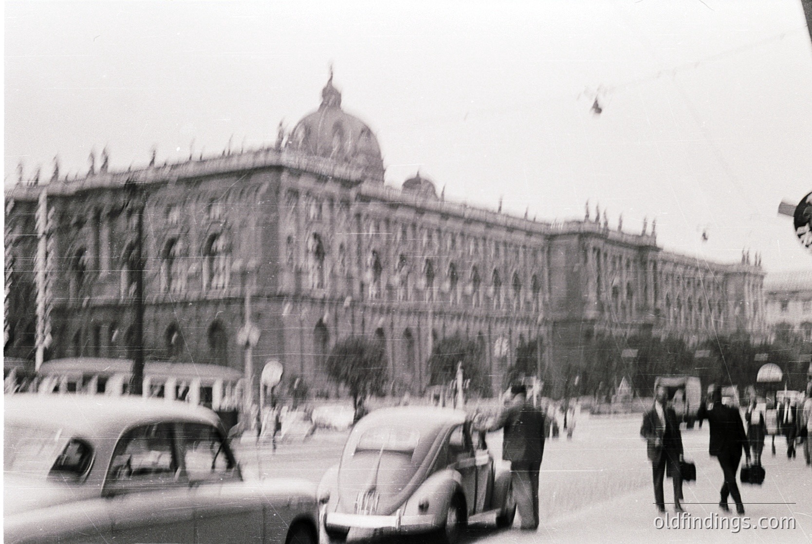 Vienna’s grand **Hofburg Imperial Palace** dominates this mid-20th century street scene, showcasing its Baroque façade and central dome. Classic **Volkswagen Beetle** and vintage cars navigate a bustling urban square, while pedestrians in 1950s-60s attire add historical context. The architectural grandeur and mid-century urban life blend seamlessly.