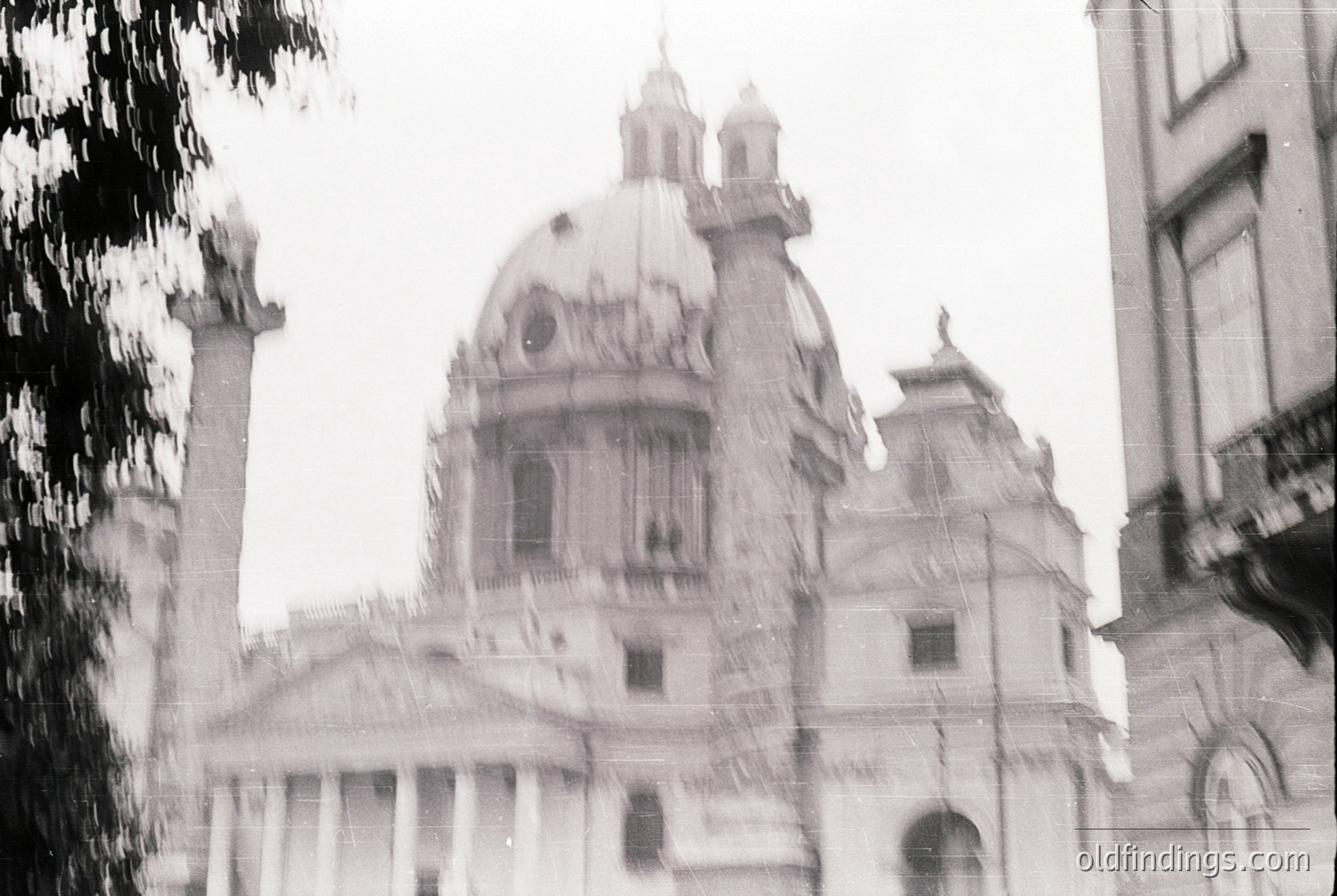 Neoclassical church dome with intricate detailing, flanked by bell towers and arched windows. Blurred foreground suggests motion or vintage film grain. Likely European urban setting, 19th–early 20th century.