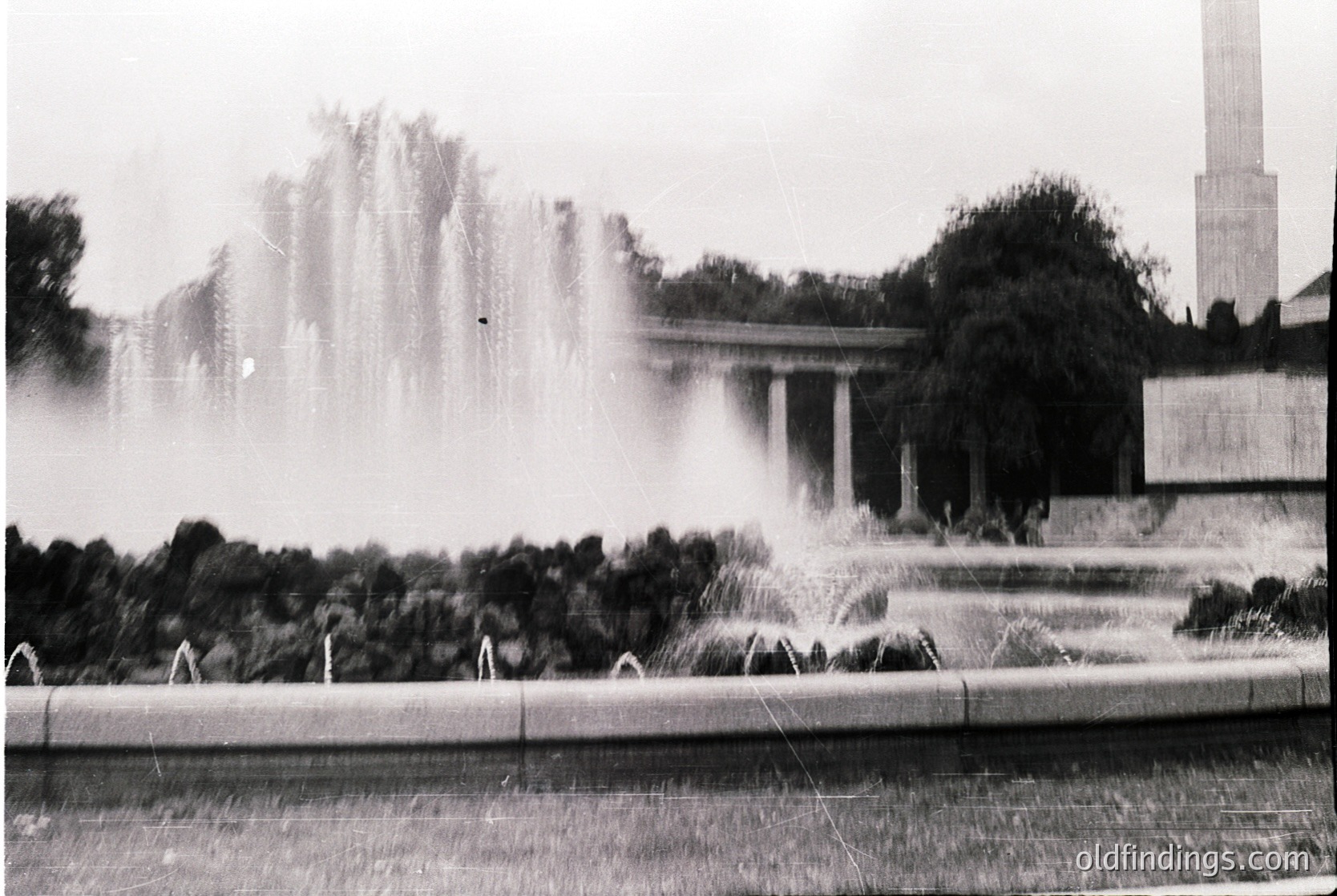 Classic black-and-white shot of a grand **fountain** in an urban plaza, framed by **classical columns** and **monumental trees**. Likely **1950s–1970s** Soviet-era architecture, featuring **neoclassical influences**. Water cascades symmetrically, creating a dynamic centerpiece.