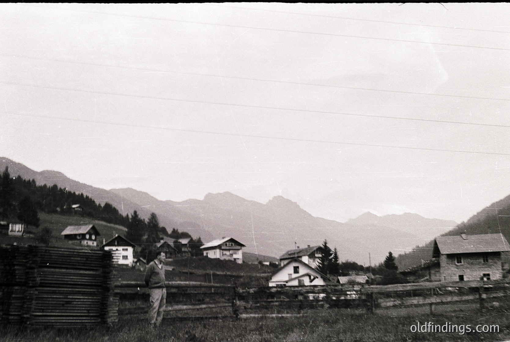 Vintage black-and-white alpine village scene with wooden chalets and rustic fencing. A man in mid-20th-century attire poses near a wooden fence, surrounded by rolling hills and dense forest.