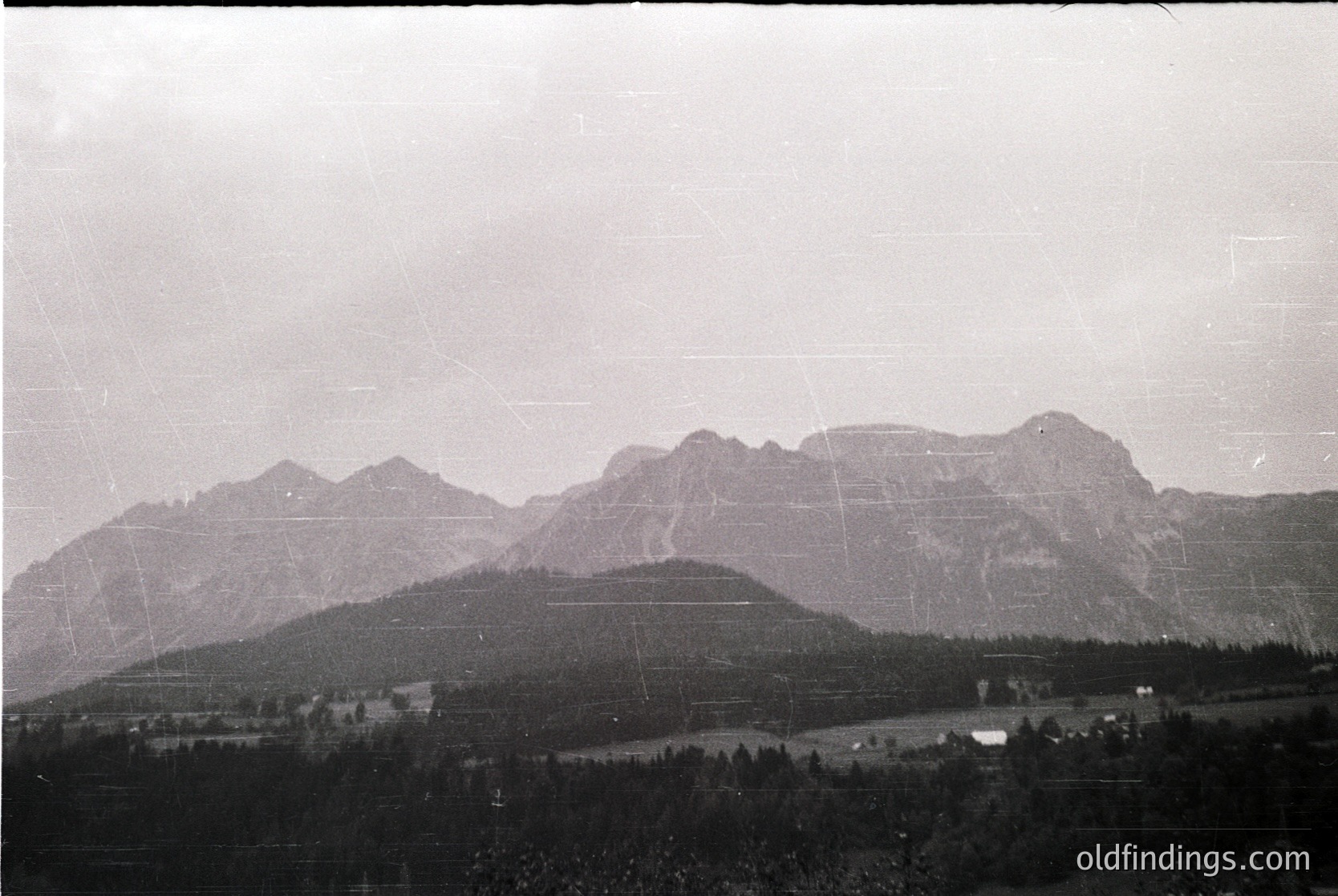 Black-and-white aerial view of rugged alpine peaks with sparse forest cover, likely mid-20th century. Distinctive jagged ridges and layered rock formations dominate the landscape. Scattered buildings and roads suggest remote settlement.
