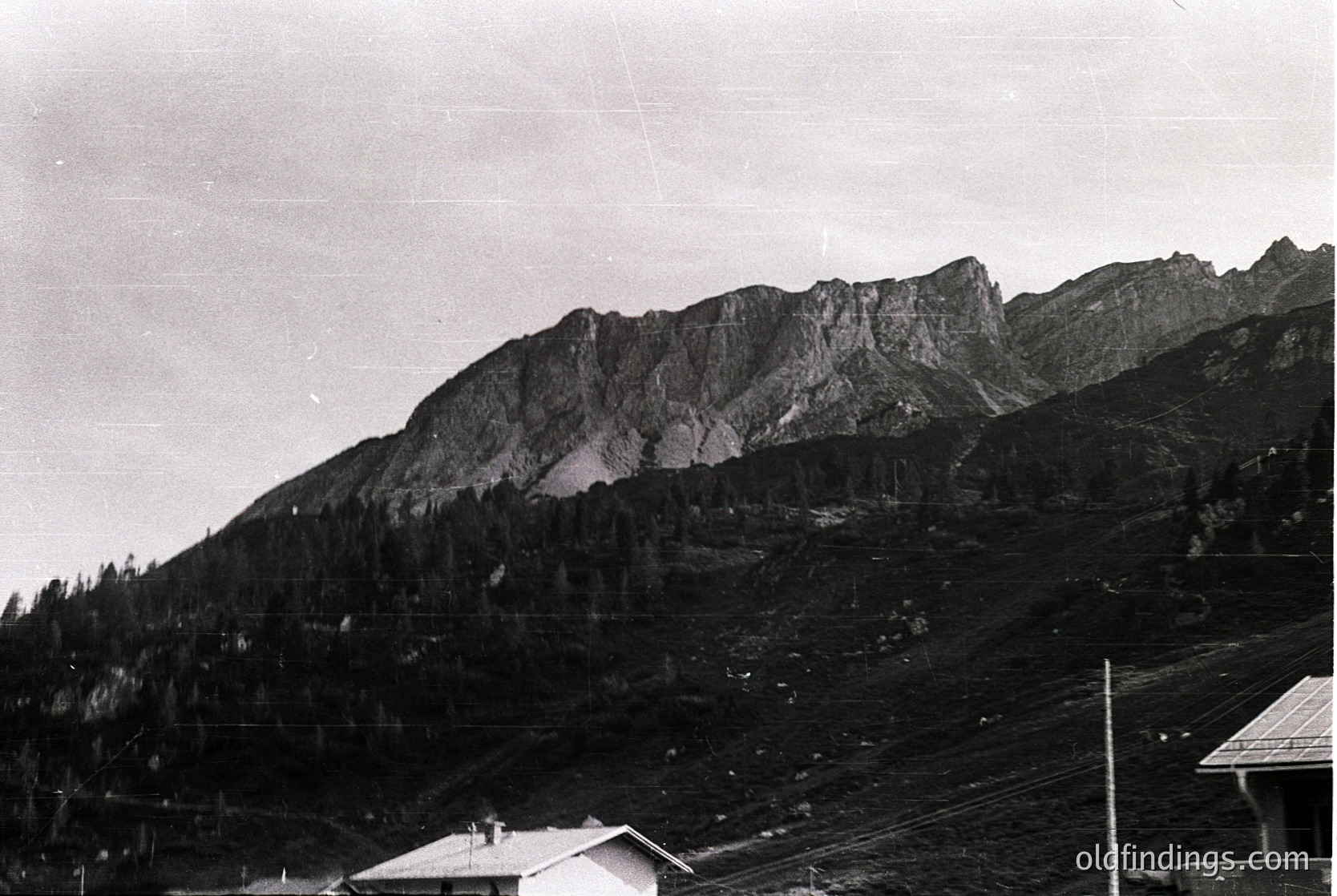 Black-and-white alpine landscape featuring jagged peaks and dense coniferous forest. Mid-20th century mountain resort architecture visible at base. Snow patches and rocky terrain dominate the midground.