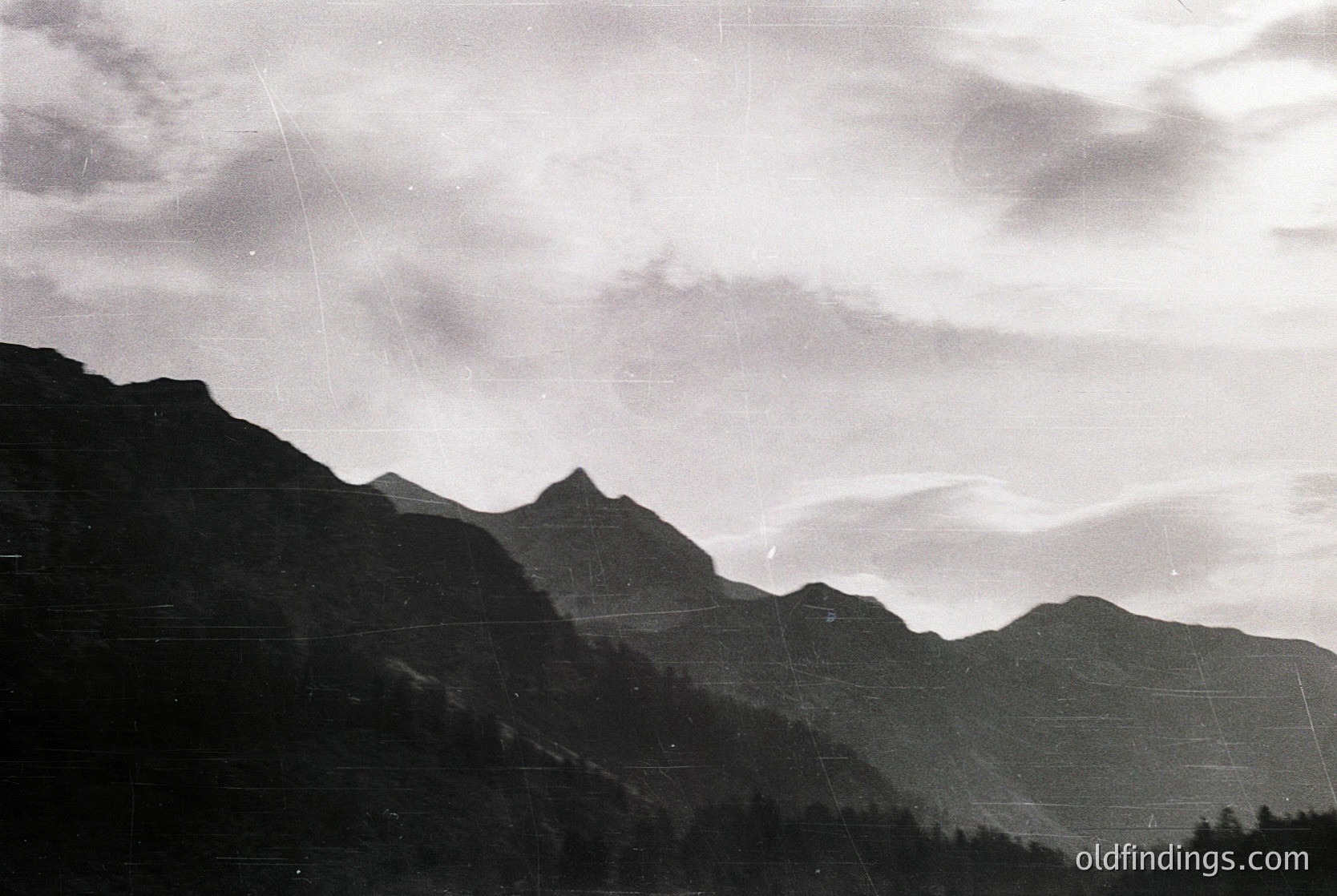 Black-and-white aerial view of rugged alpine peaks with dense forest cover, likely captured mid-20th century. Dramatic cloud formations contrast with jagged ridges, suggesting high-altitude terrain. Potential research value for geology or historical aviation imagery.