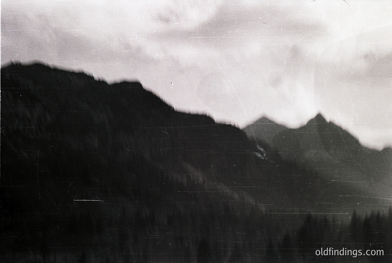 Vintage black-and-white mountain landscape with dense forest cover and misty peaks, evoking a moody, atmospheric feel. Likely mid-20th century due to grainy texture and monochrome quality.