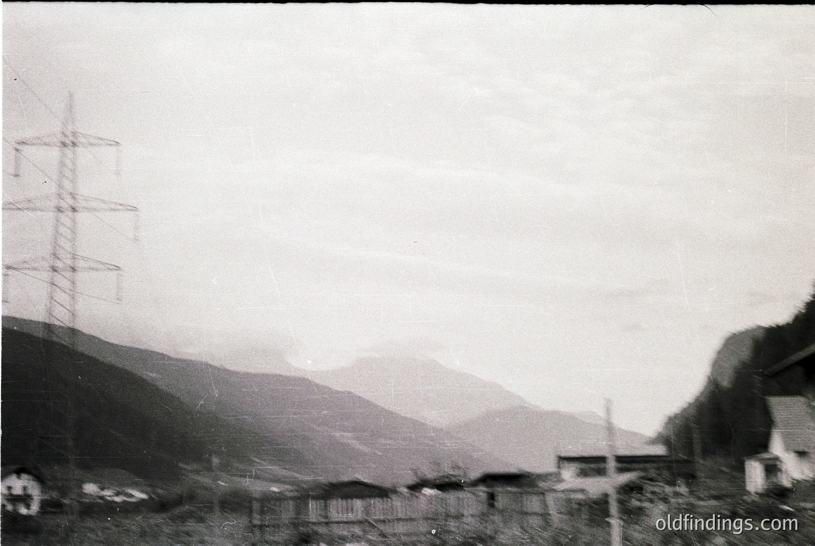 Black-and-white rural landscape featuring a high-voltage pylon on left, spanning across a misty valley. Low-lying buildings and scattered structures in foreground, with rolling hills and mountains in background. Likely Eastern European, mid-20th century.