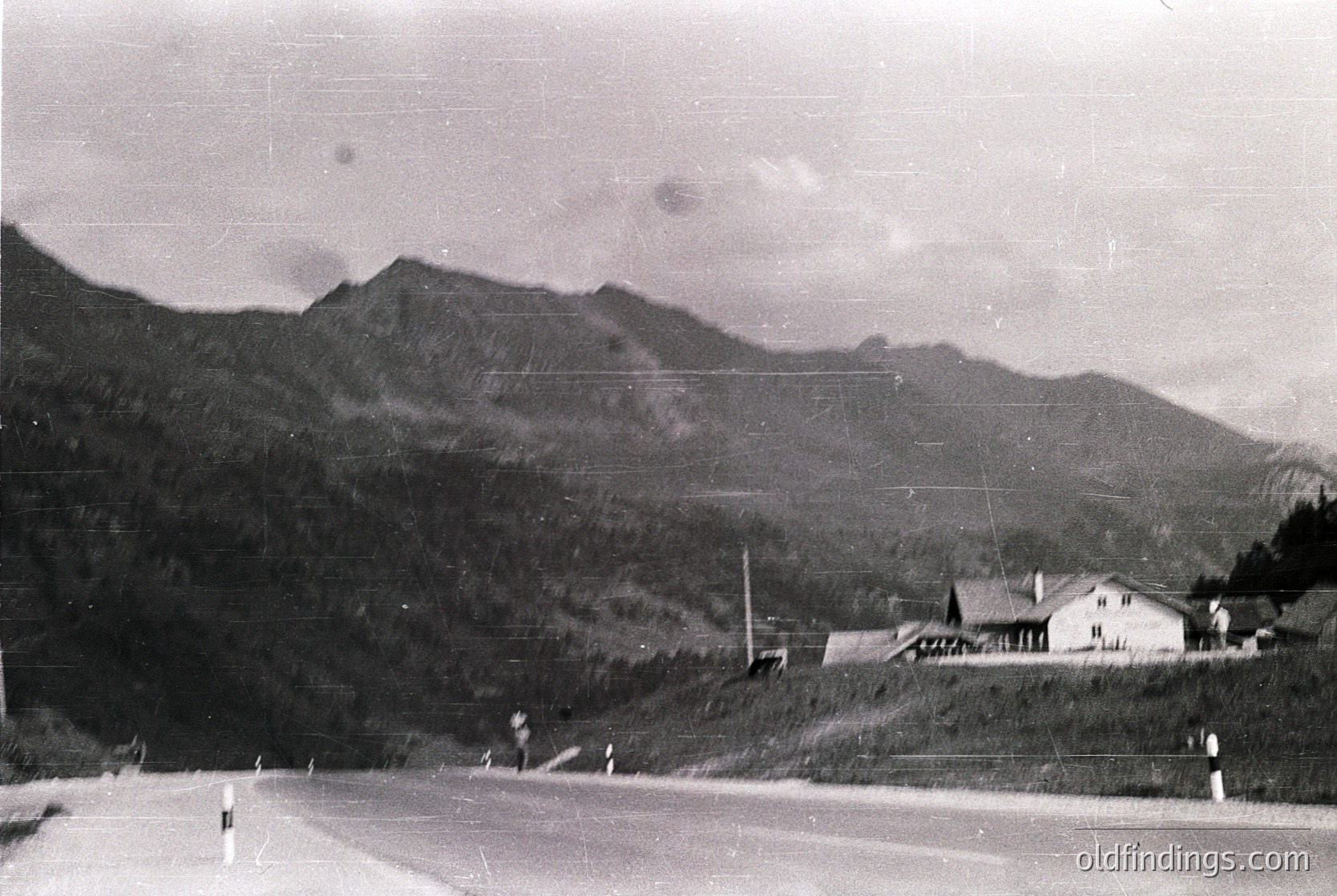 Mid-20th century alpine village nestled at mountain base, featuring a single-story wooden house with pitched roof. Road curves left, bordered by wooden posts. Dense forested slopes rise steeply behind.