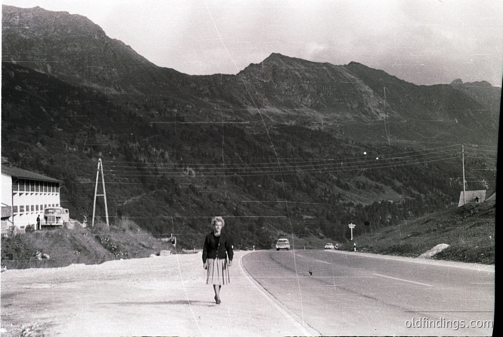 Mid-20th century alpine roadside scene: woman in 1950s-style dress crossing a winding mountain road flanked by utility poles. Snow-capped peaks and forested slopes in background. Single-story building with balcony at left. Vintage cars parked along road.